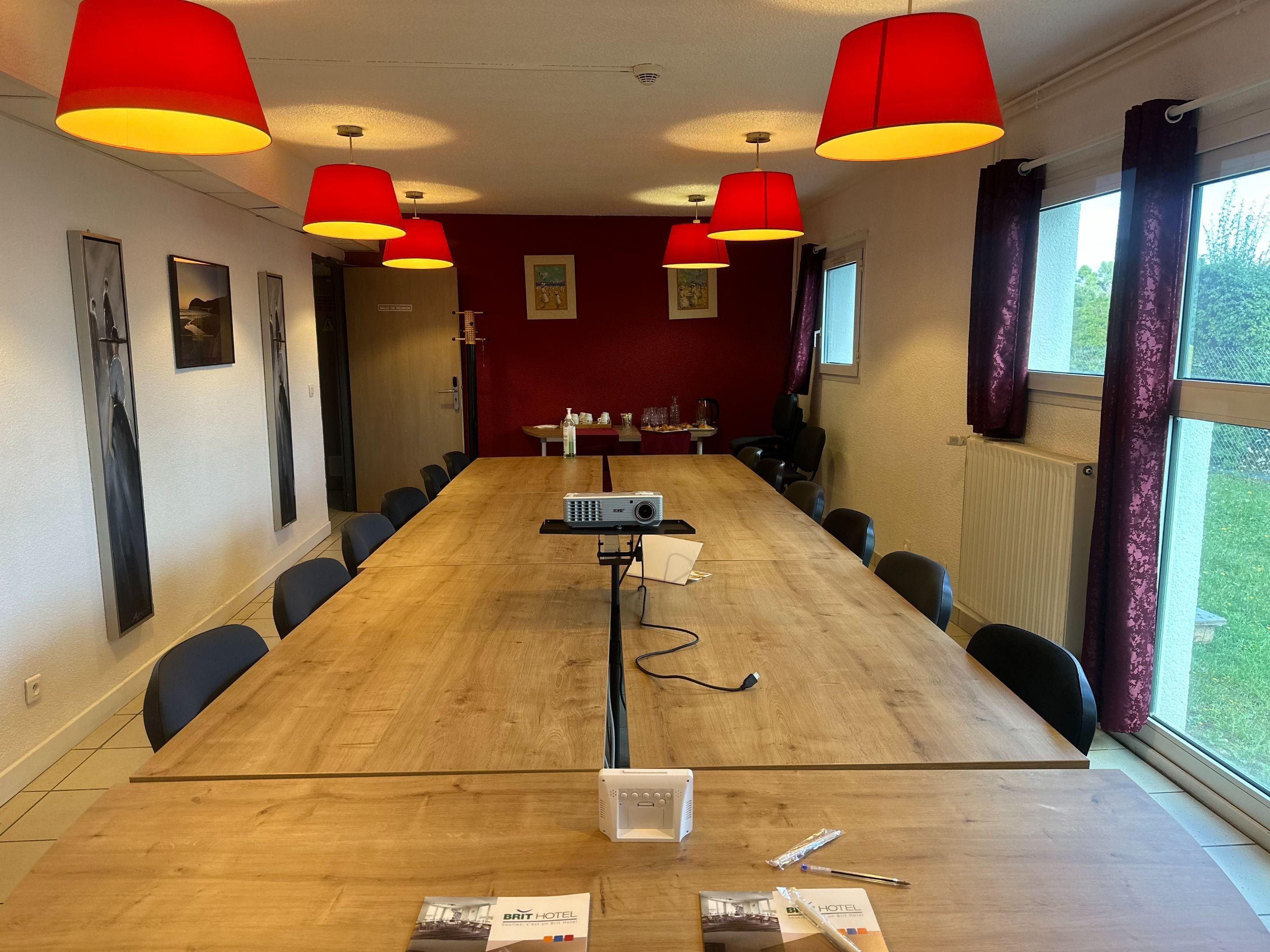 Una sala conferenze con un lungo tavolo, sedie, un proiettore e lampadari a sospensione rossi.