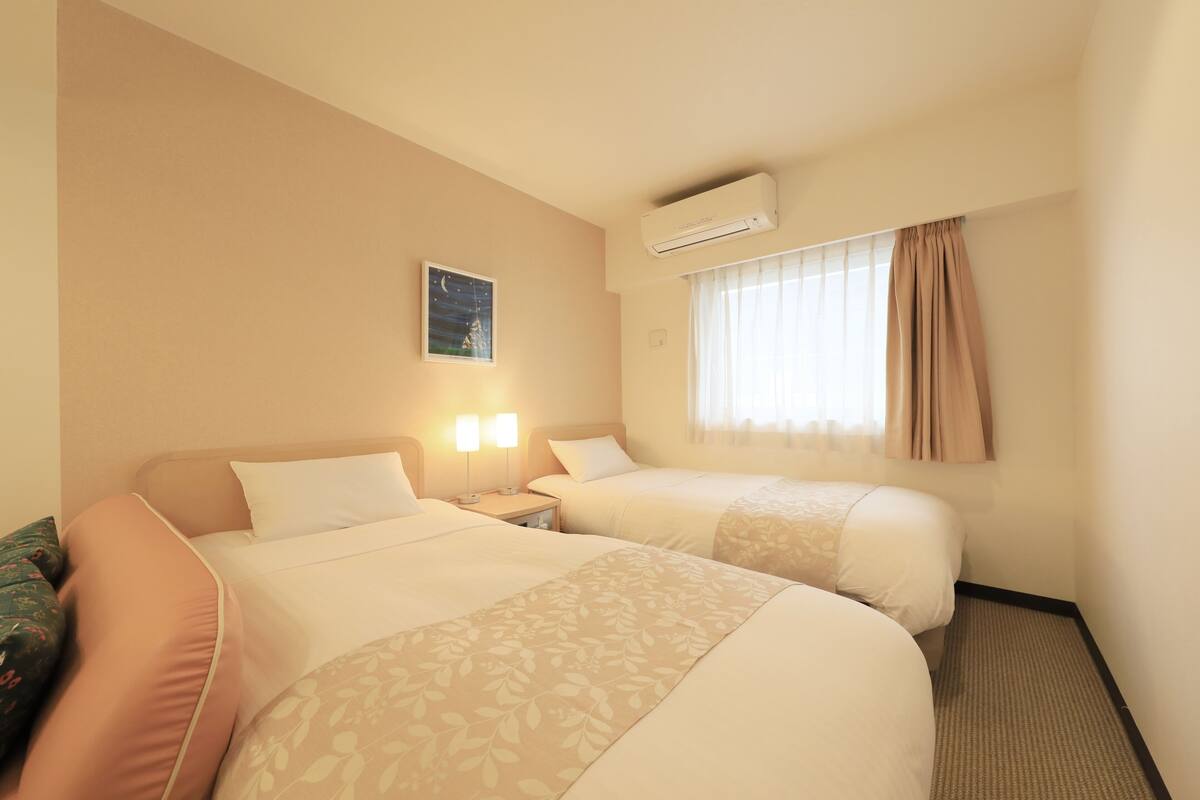 Une chambre d’hôtel avec deux lits, une table de chevet avec des lampes, un climatiseur, une fenêtre avec des rideaux et un tableau accroché au mur.