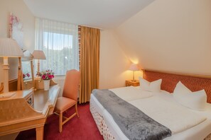 Une chambre d’hôtel avec un lit, un bureau, une chaise, une lampe et une fenêtre avec des rideaux.