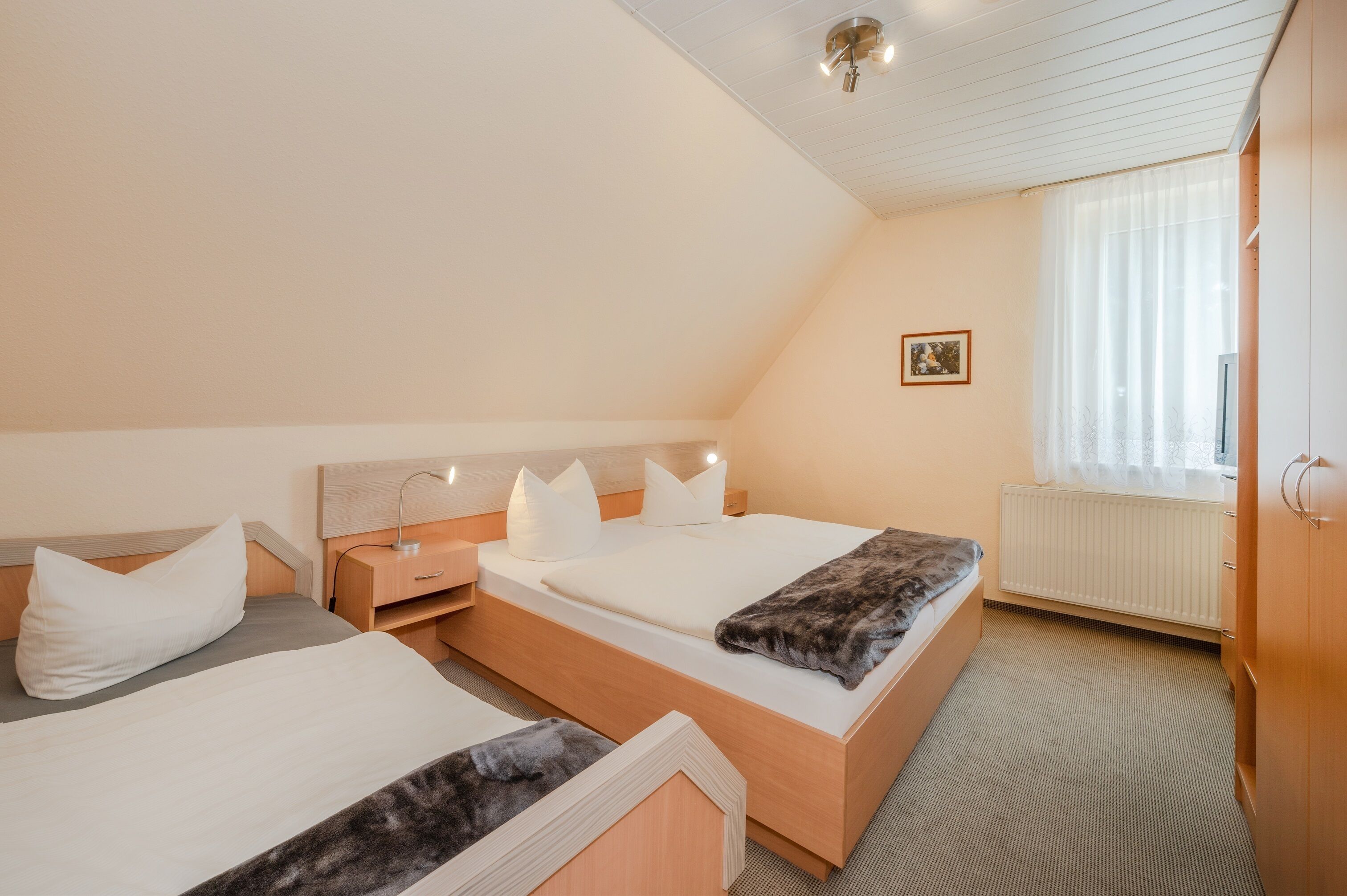 A hotel room with two beds, a wooden headboard, a bedside table with a lamp, a window with sheer curtains, and a radiator.