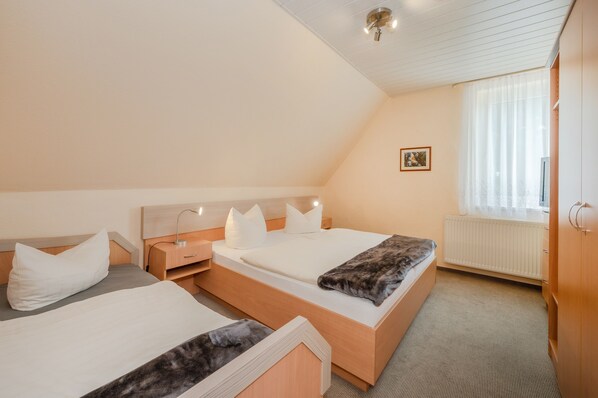 Hotellihuone, jossa on kaksi sänkyä, puinen pääty, yöpöytä lampulla, ikkuna, jossa on läpikuultavat verhot, ja patteri.