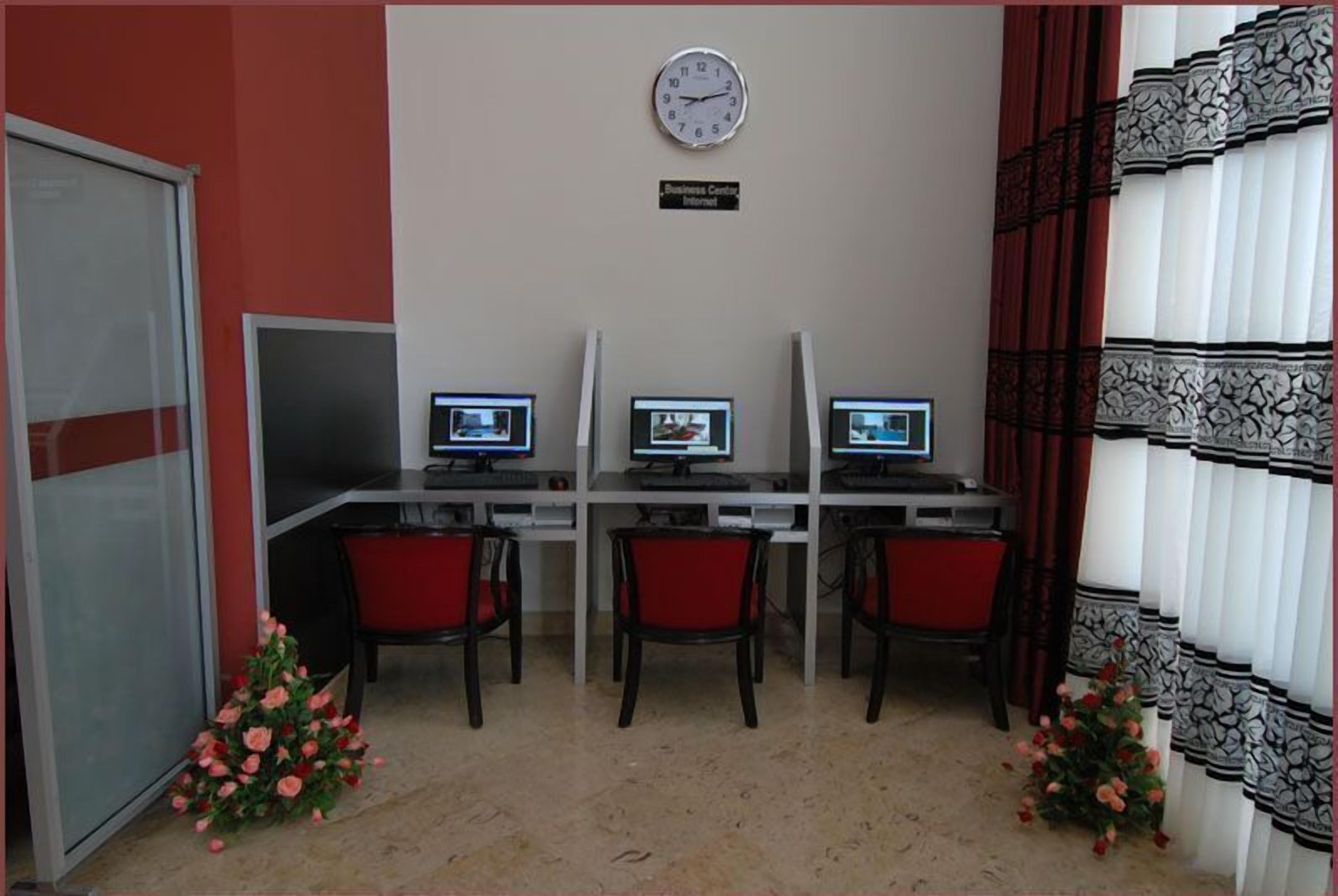 Een business center met drie computerwerkplekken, rode stoelen, een klok en decoratieve bloemen.
