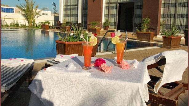 A poolside area with lounge chairs, a table with drinks, and a view of a building with large windows. - Appart Hotel Founty Beach (Agadir)