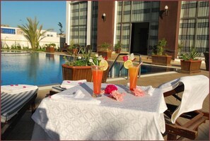 Un espace aménagé au bord de la piscine, avec des chaises longues, une table où sont servis des boissons et une vue sur un bâtiment doté de grandes fenêtres.