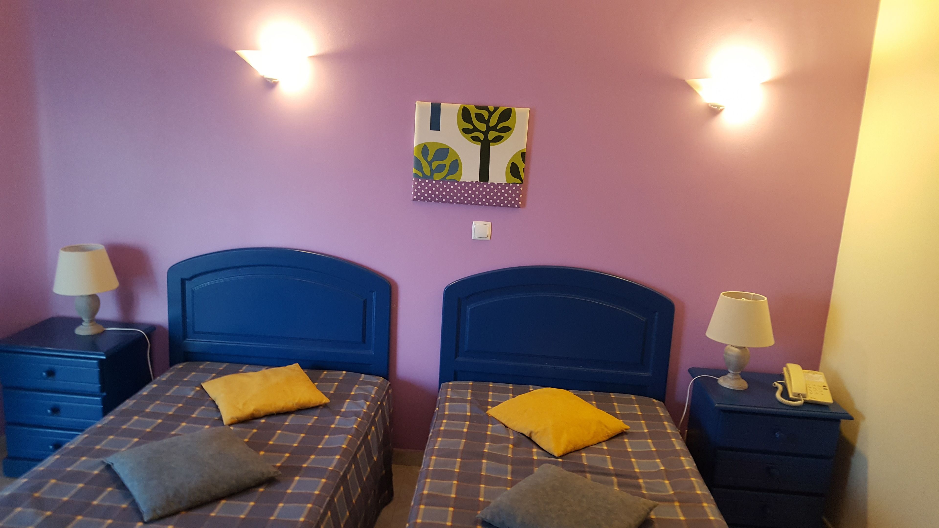 A room with two beds, each with a headboard and a bedside table with a lamp. There is a wall-mounted light fixture above the beds and a small wall decoration.