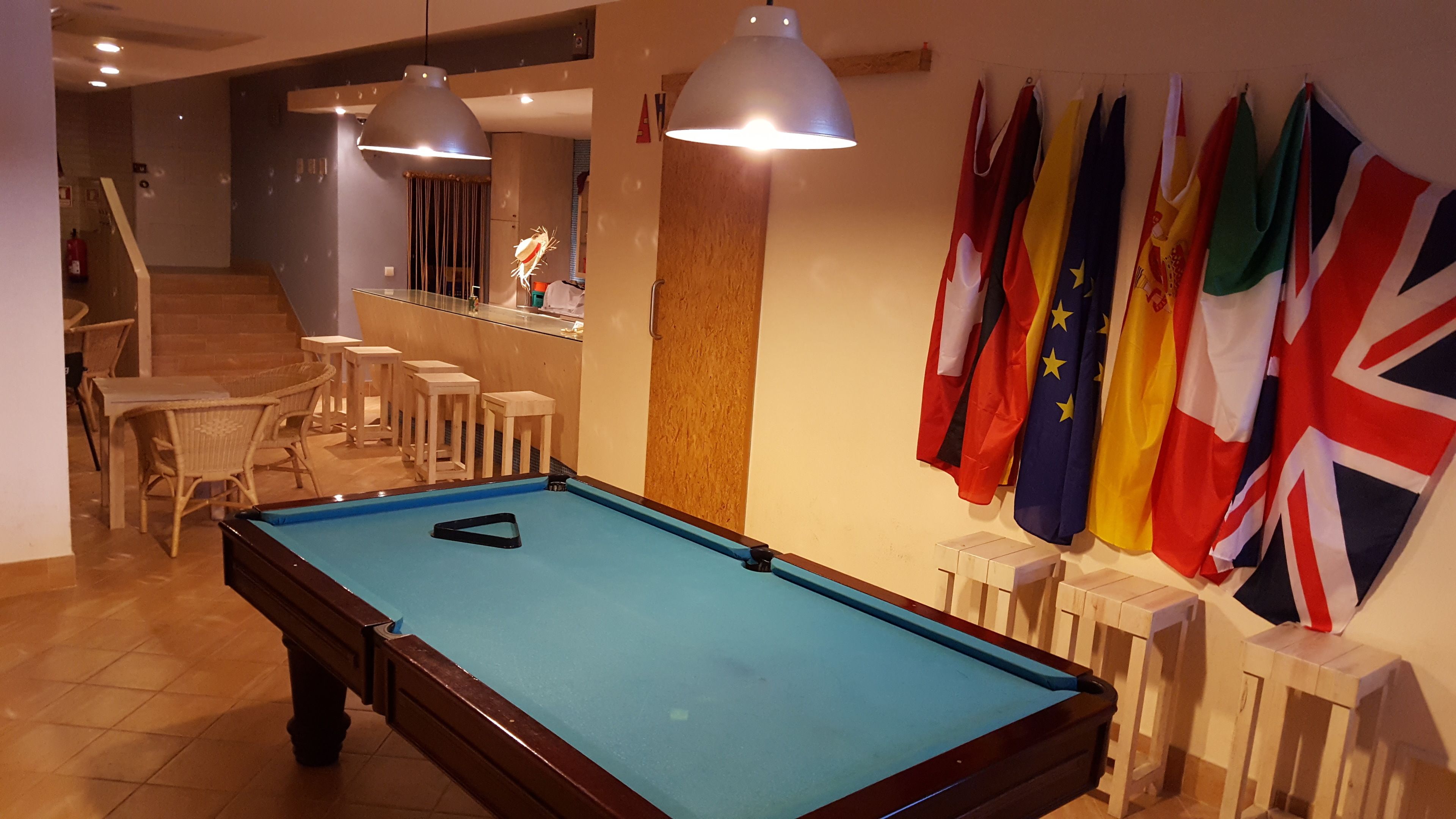 Una mesa de billar con superficie azul de fieltro, rodeada de banderas, una zona de bar con taburetes y una escalera que sube a un nivel superior.