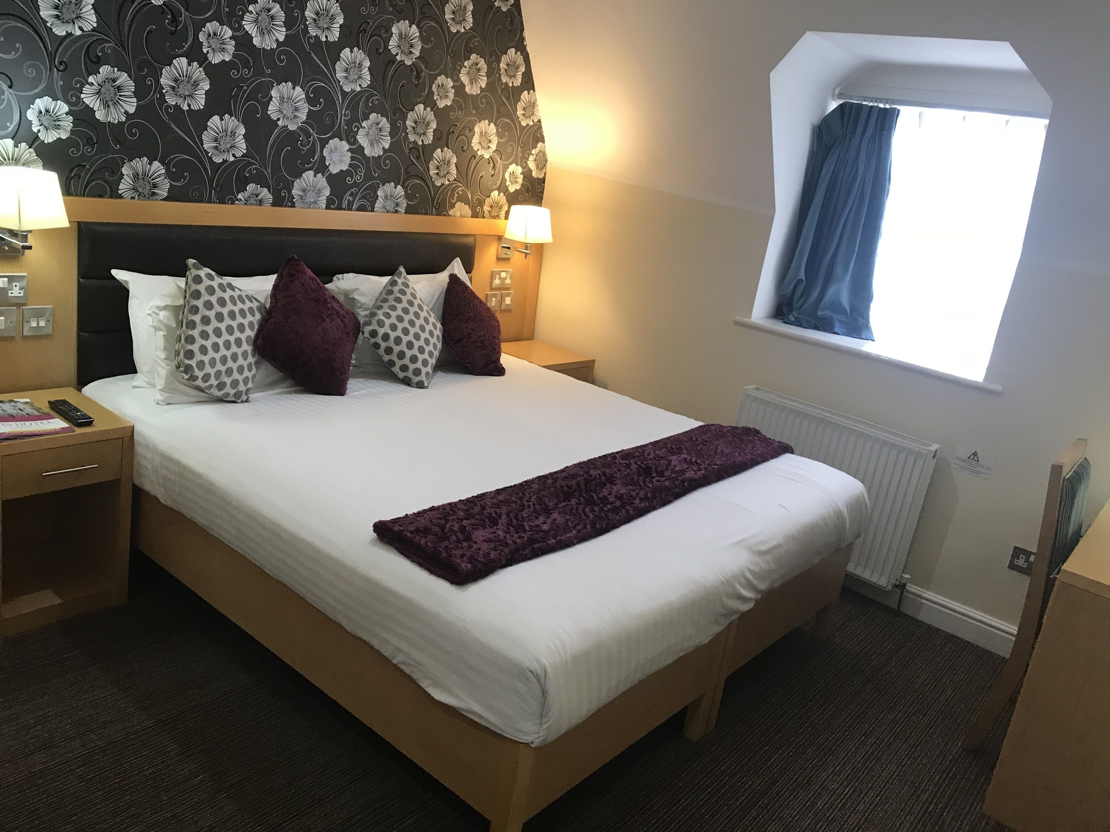 A hotel room with a large bed, a wooden headboard, a window with blue curtains, and a floral wallpaper design.