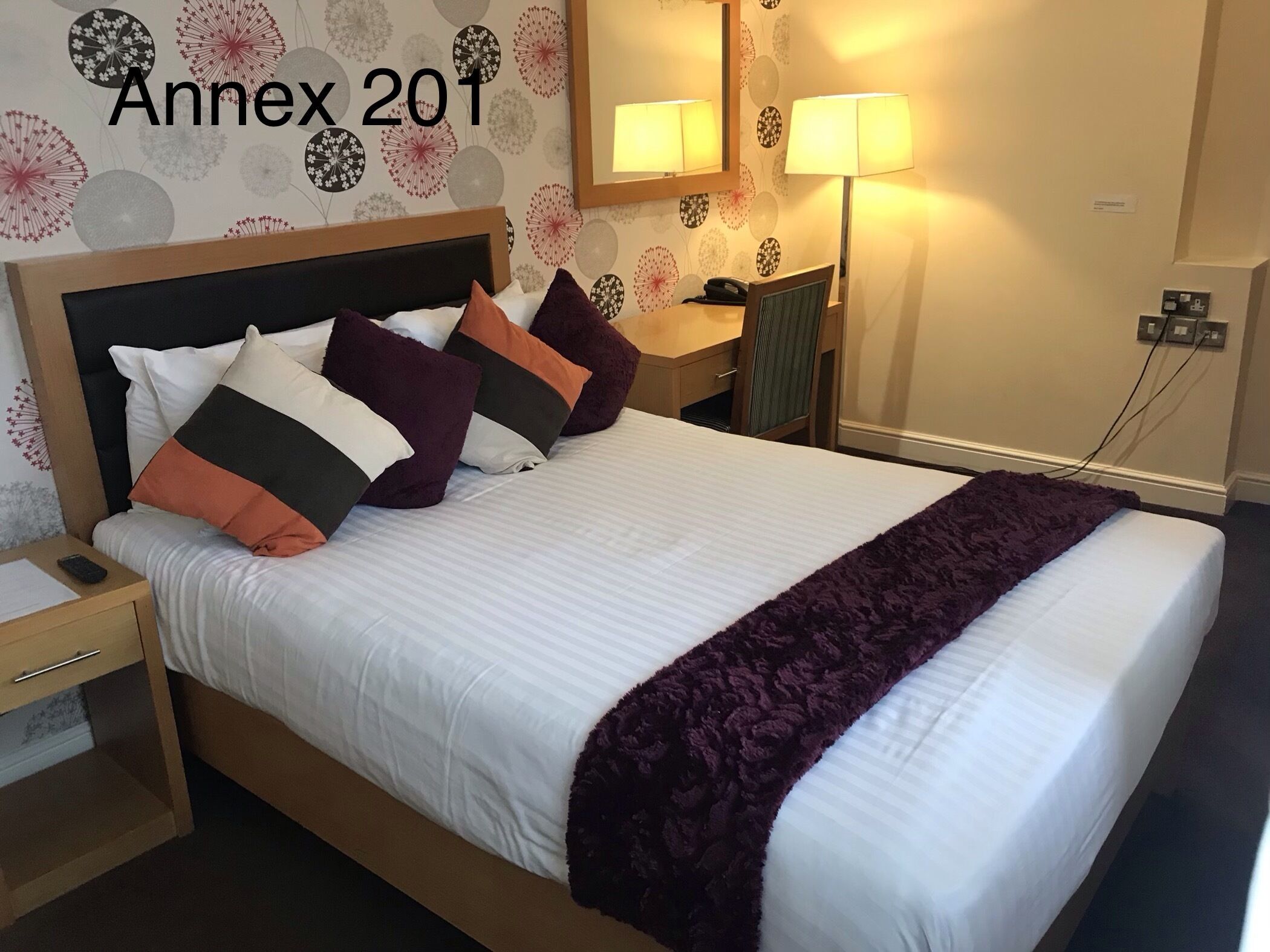 Un lit bien fait, avec une couette blanche et un plaid violet, une table de chevet avec une télécommande, et un mur orné d’un motif floral et portant l’inscription « Annexe 201 ».