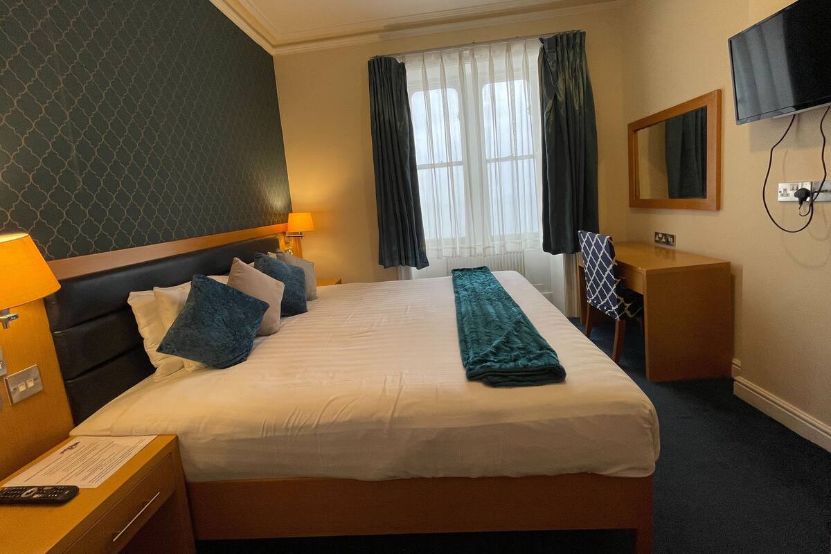 A hotel room with a bed, two bedside lamps, a desk, a chair, a television, and a window with curtains.