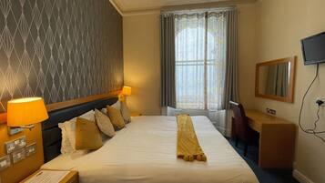 A hotel room with a neatly made bed, two bedside lamps, a desk with a chair, a television mounted on the wall, and a window with curtains.
