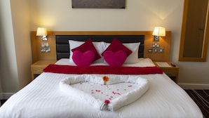 Ein ordentlich bezogenes Bett mit einer Kissenanordnung in Herzform, einer roten Tagesdecke und einer floralen Tischdekoration.