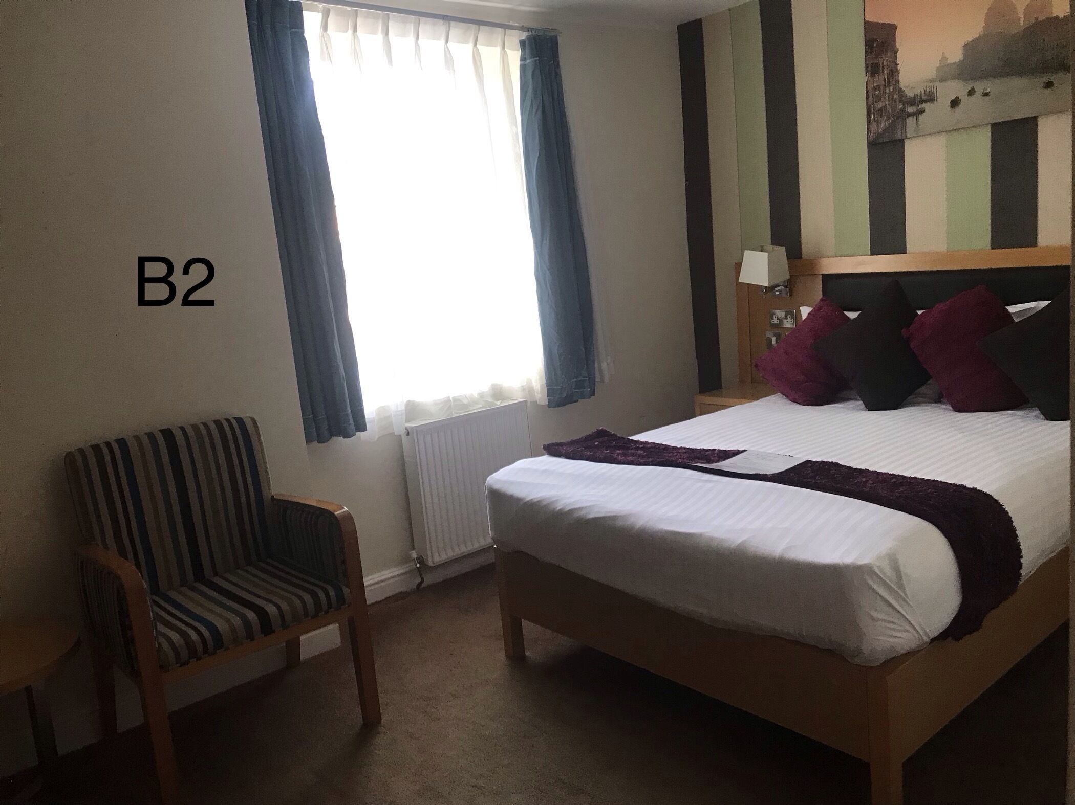 A hotel room with a bed, a chair, a window with curtains, a radiator, and a painting on the wall.