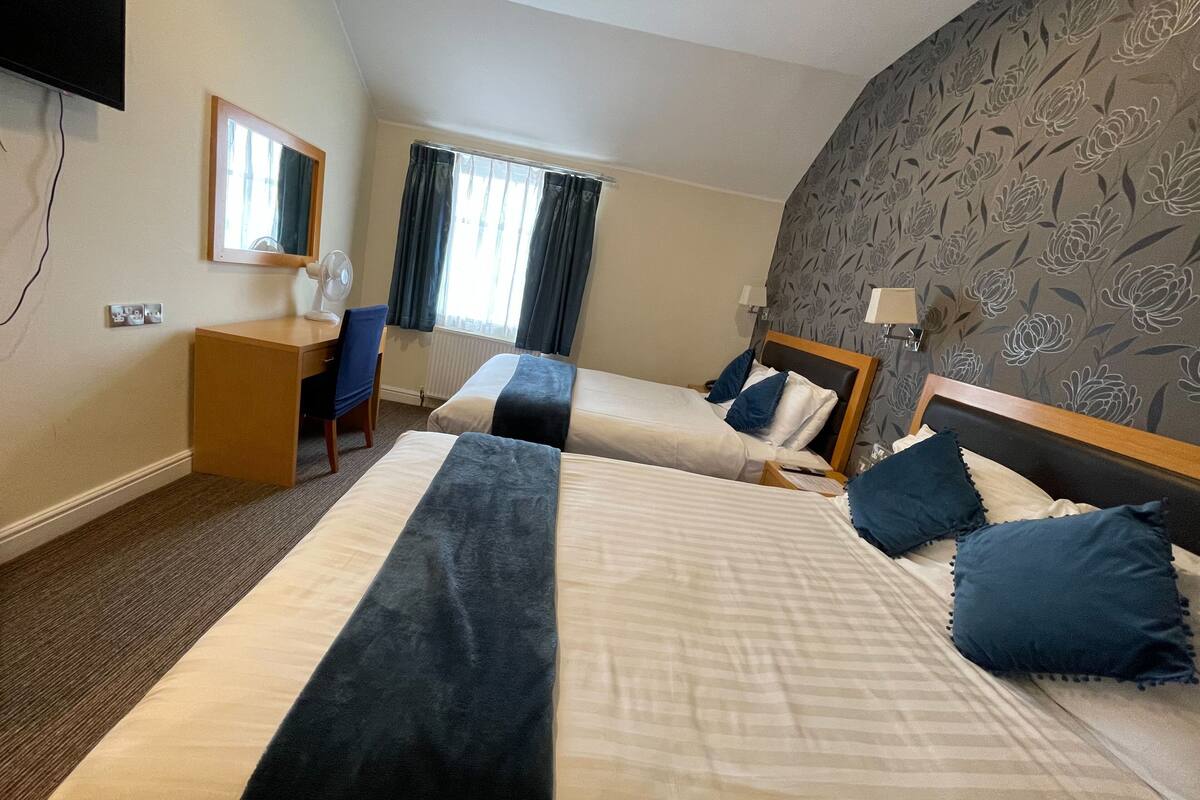 A hotel room with two beds, a desk, a chair, a window with curtains, and a wall with floral wallpaper.