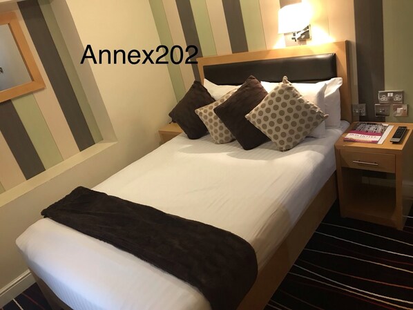 A hotel room with a bed, pillows, a bedside table with a lamp, a book, and a power outlet.