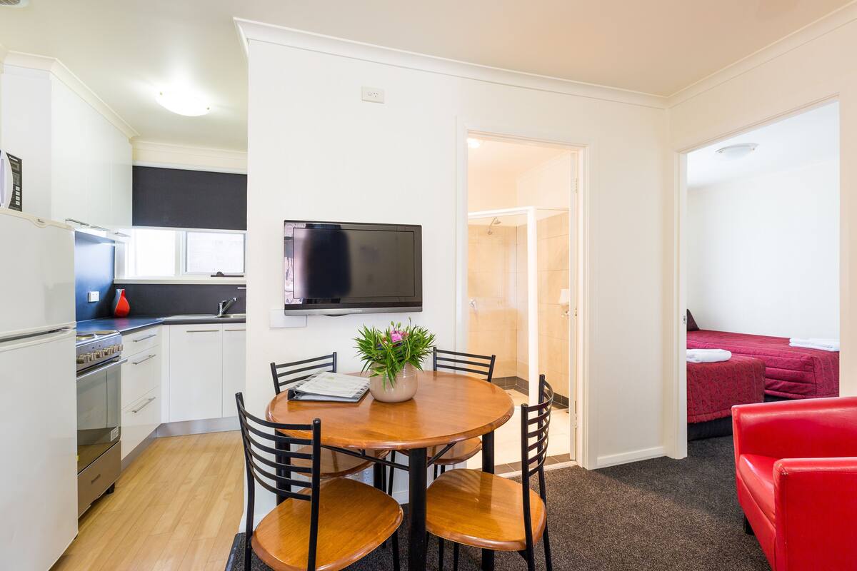 A compact living space with a kitchenette, a dining area with a table and chairs, a TV mounted on the wall, and a red couch in the living area.