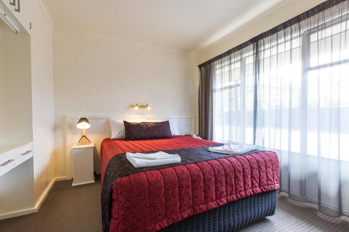 A hotel room with a bed, a nightstand with a lamp, a window with sheer curtains, and a wall-mounted light fixture.