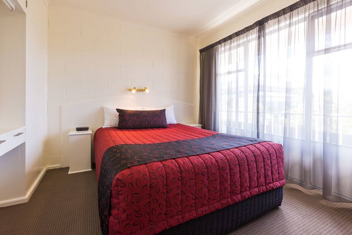 A hotel room with a bed, a nightstand, a wall-mounted light, and a window with sheer curtains.