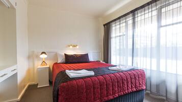 A neatly made bed with a red and black patterned bedspread, a white bedside lamp, and a window with sheer curtains.