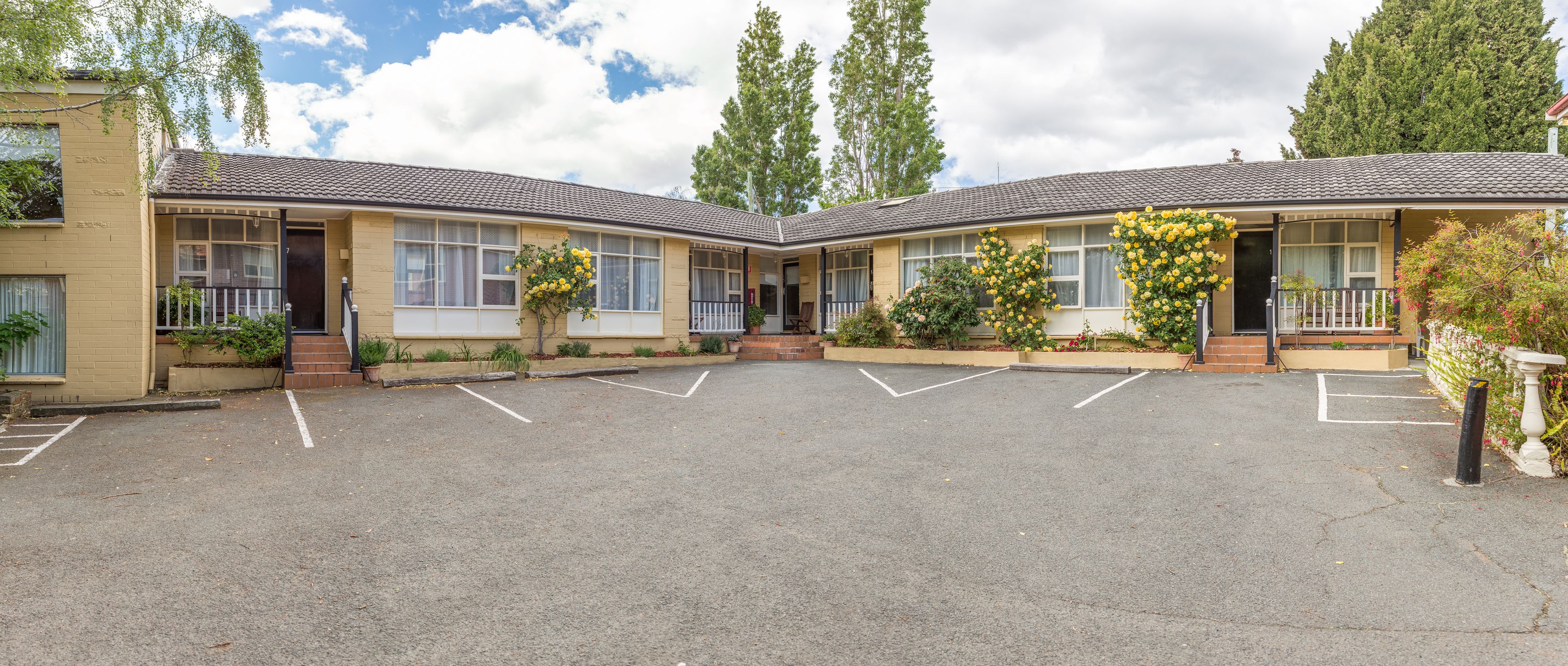 A motel with two-story buildings, a parking lot, and a garden area with yellow flowers.