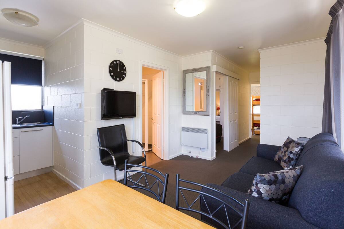 A compact living space with a kitchenette, a dining area with a table and chairs, a sofa with decorative pillows, a flat-screen TV mounted on the wall, and a mirror above the door.