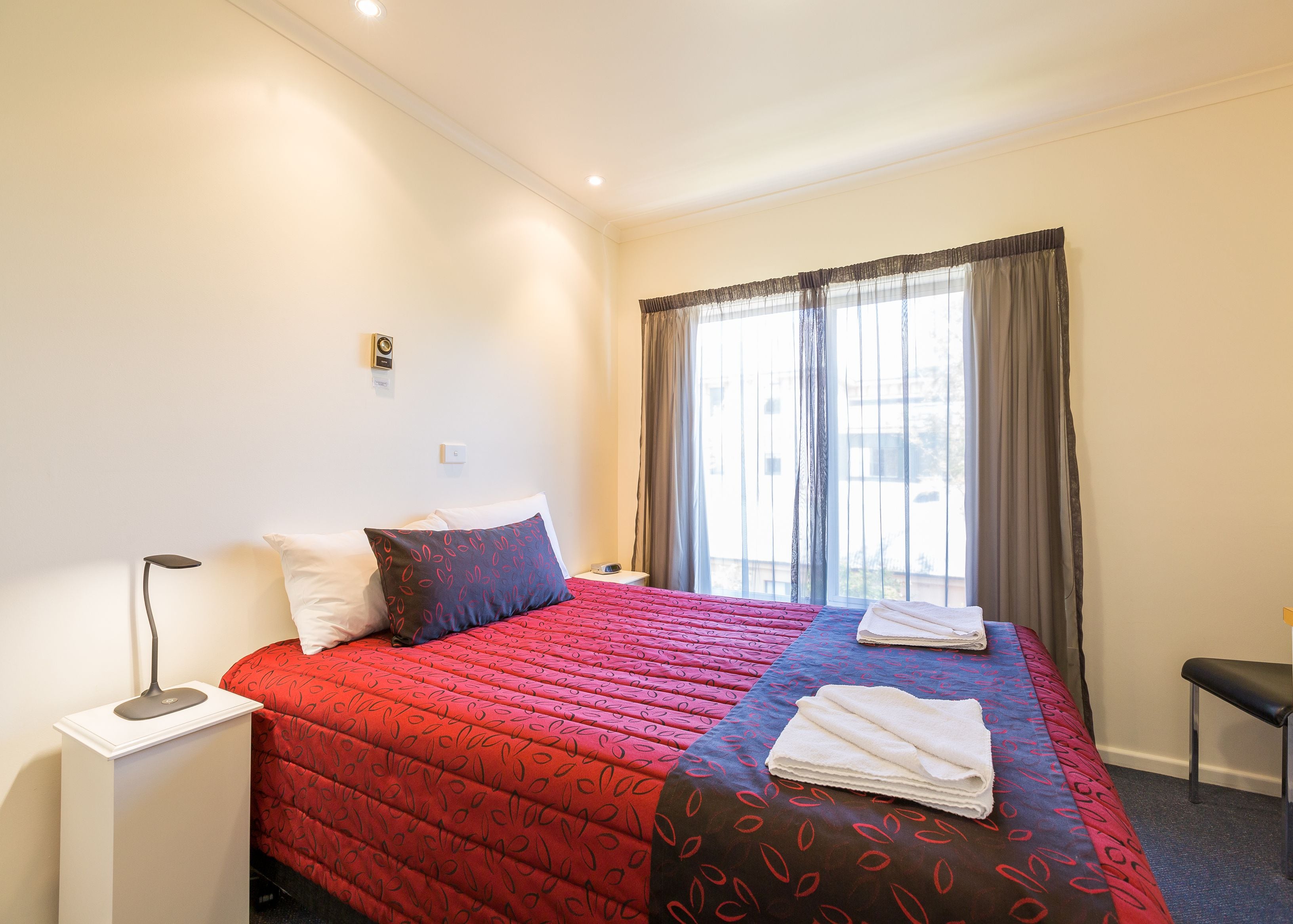 A hotel room with a bed, a bedside table, a lamp, a window with curtains, and a view of the outside.