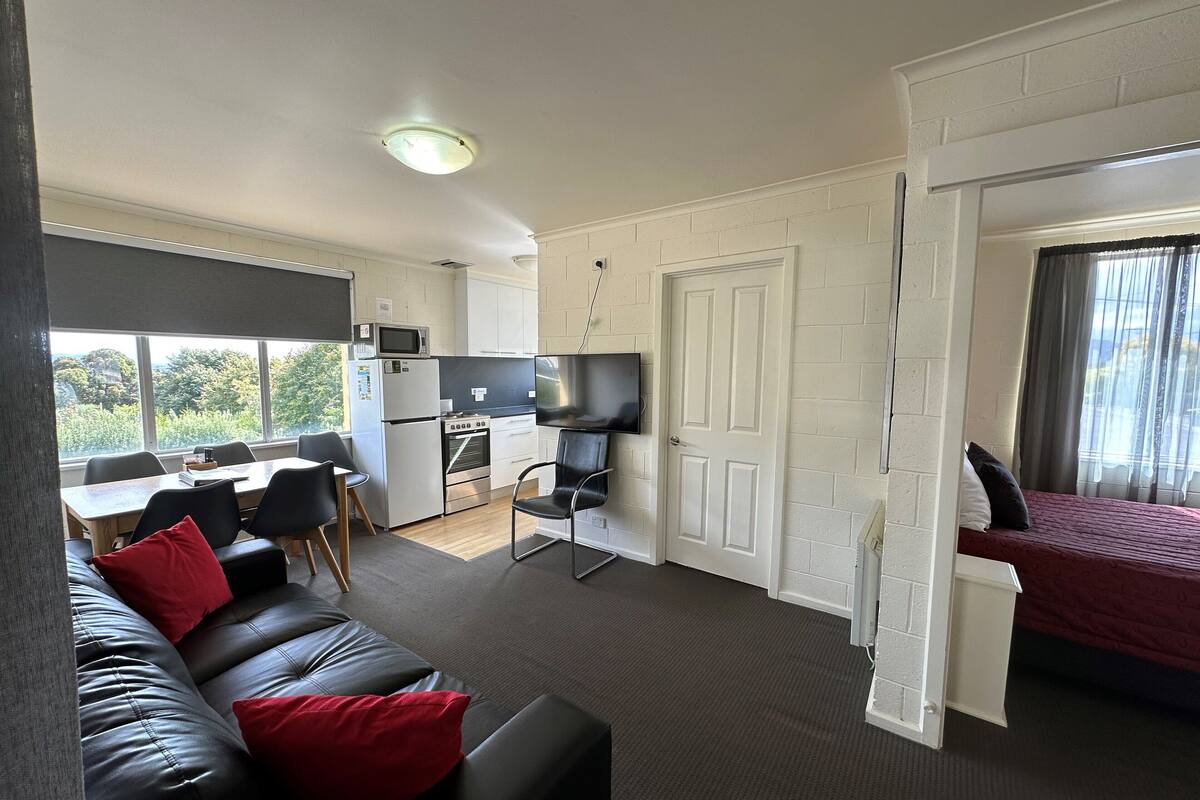 A compact living space with a kitchenette, a dining area, a sofa with red cushions, and a bed with a red bedspread.