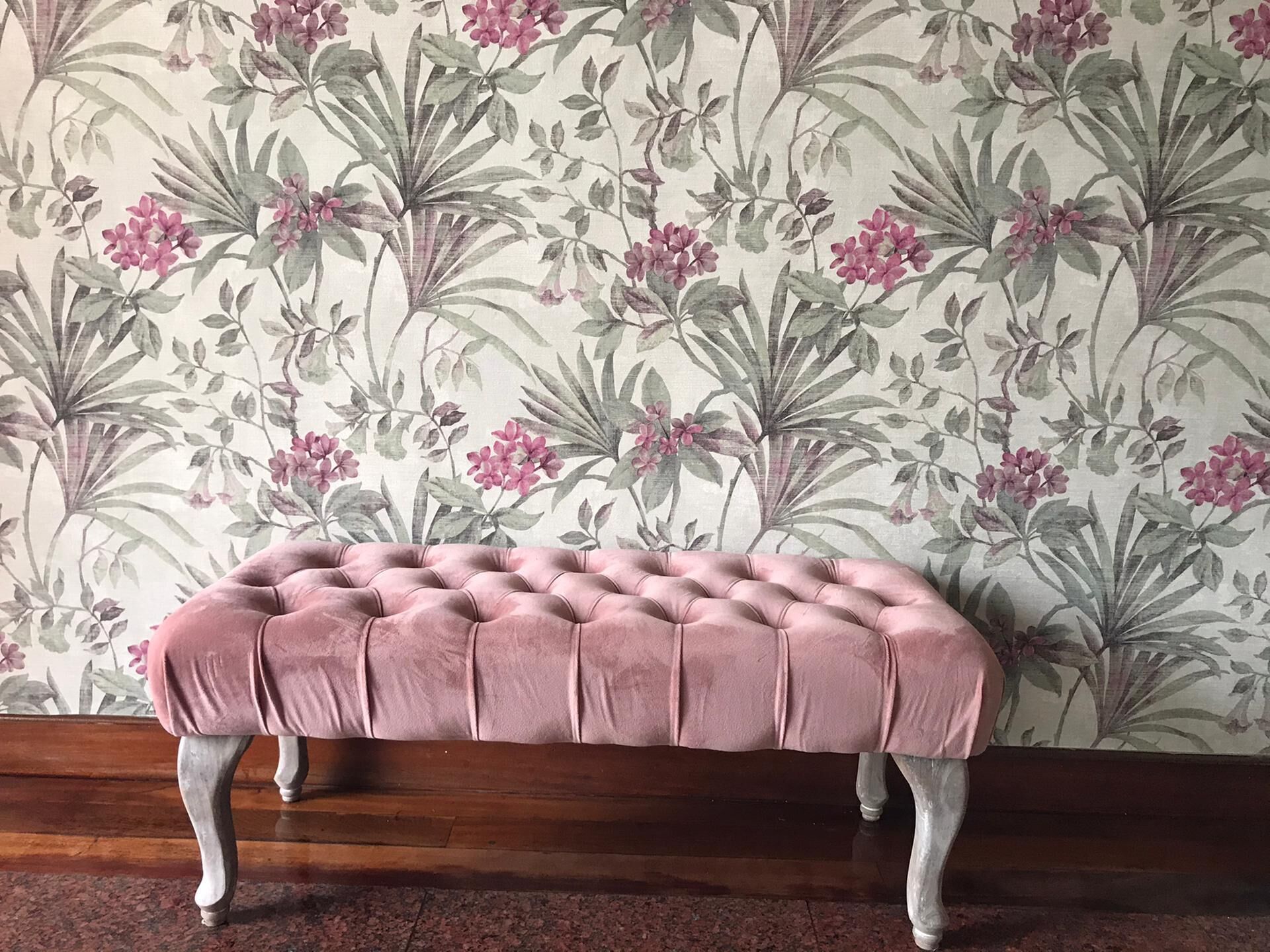 Un banco tapizado con patas de madera frente a un papel tapiz floral.