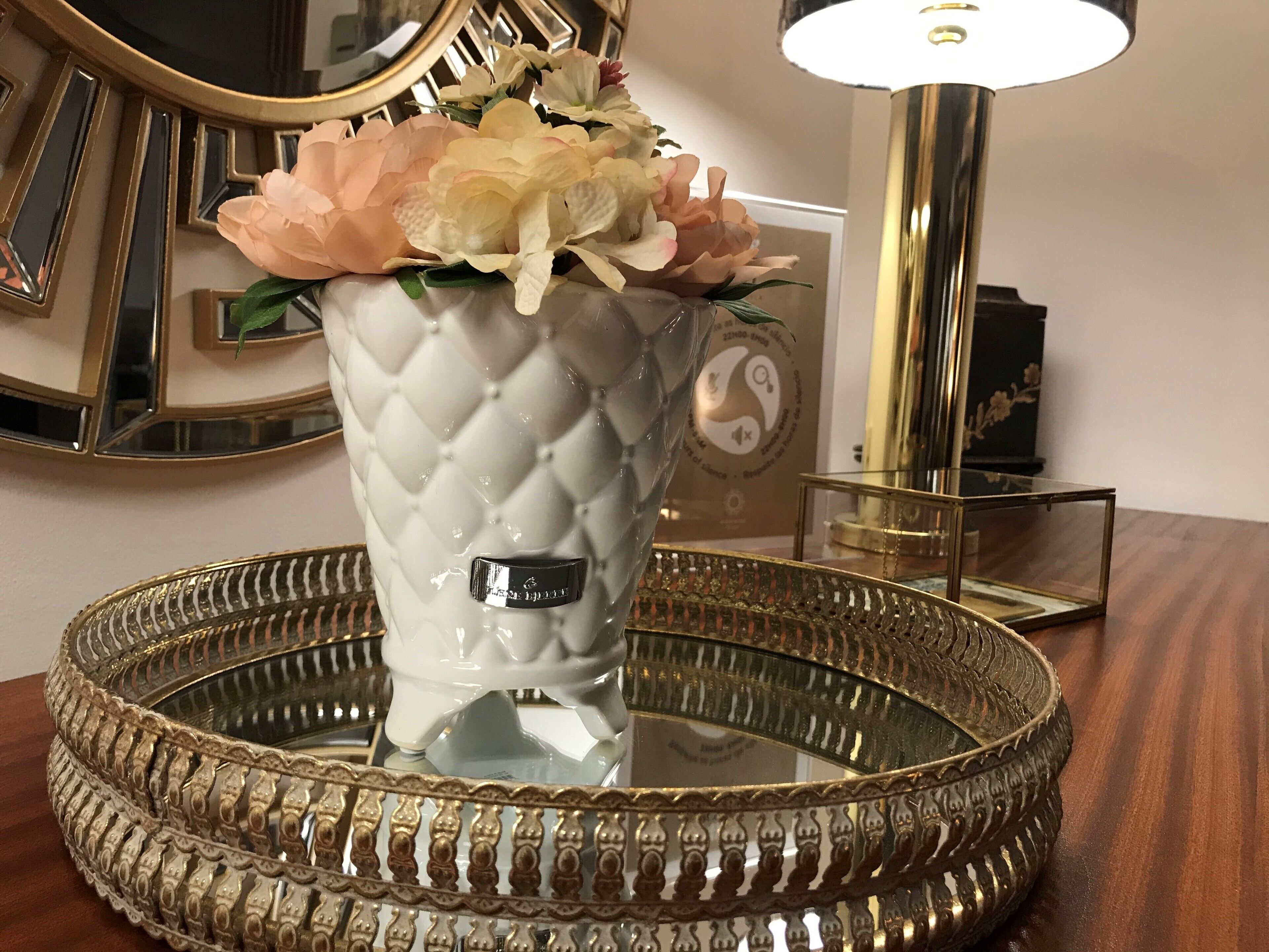 En hvid vase med et quiltet mønster og etiket, fyldt med blomster, placeret på et bakke med guld kant.