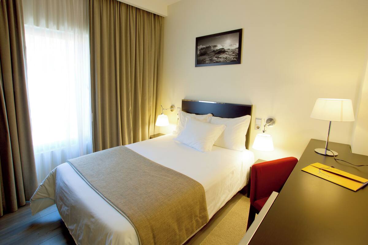 Una habitación de hotel con una cama grande, dos lámparas de noche, un escritorio con lámpara, una silla roja y un cuadro en la pared.
