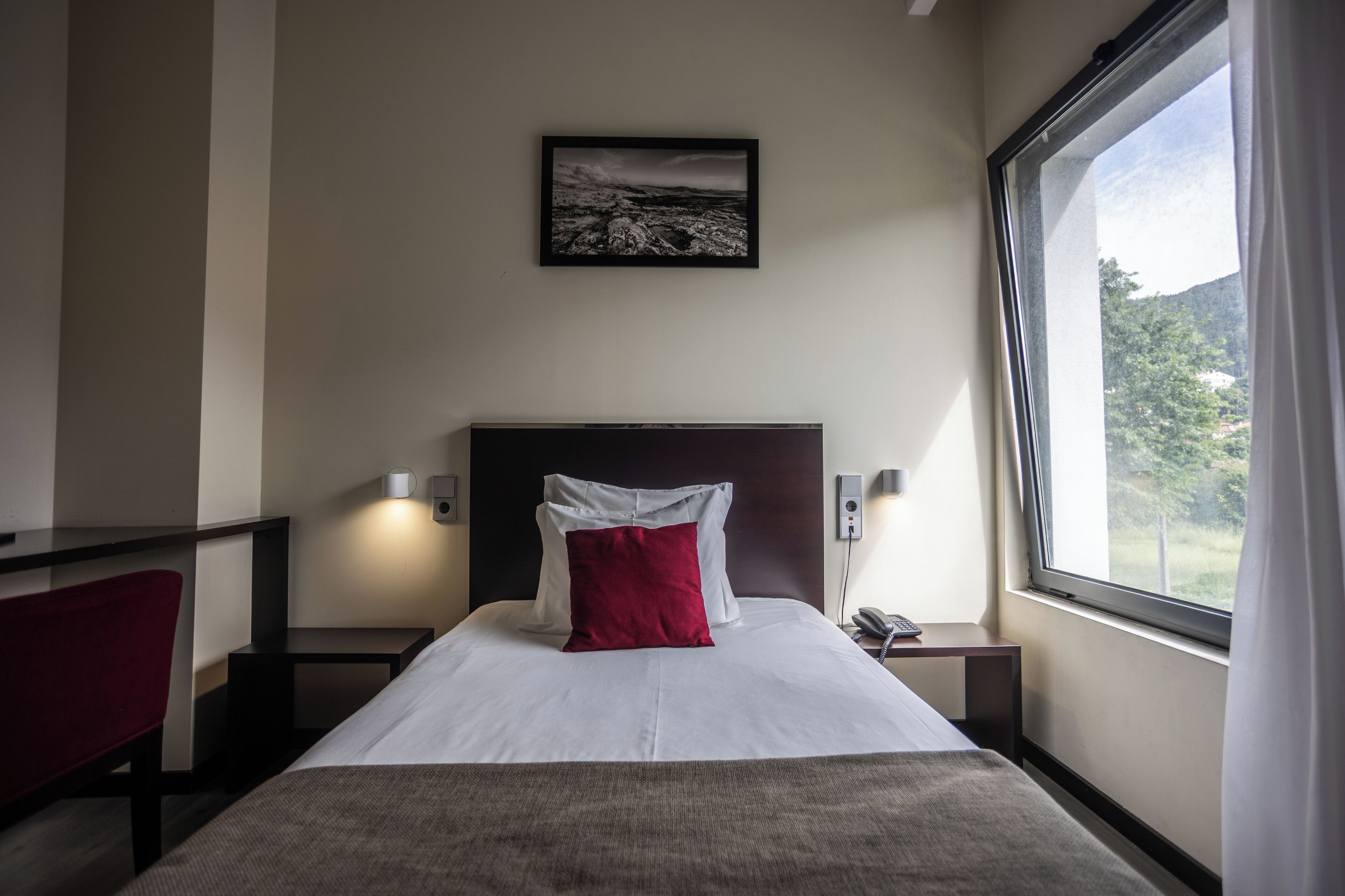 A hotel room with a bed, a nightstand, a phone, a lamp, and a window with a view of the outdoors.