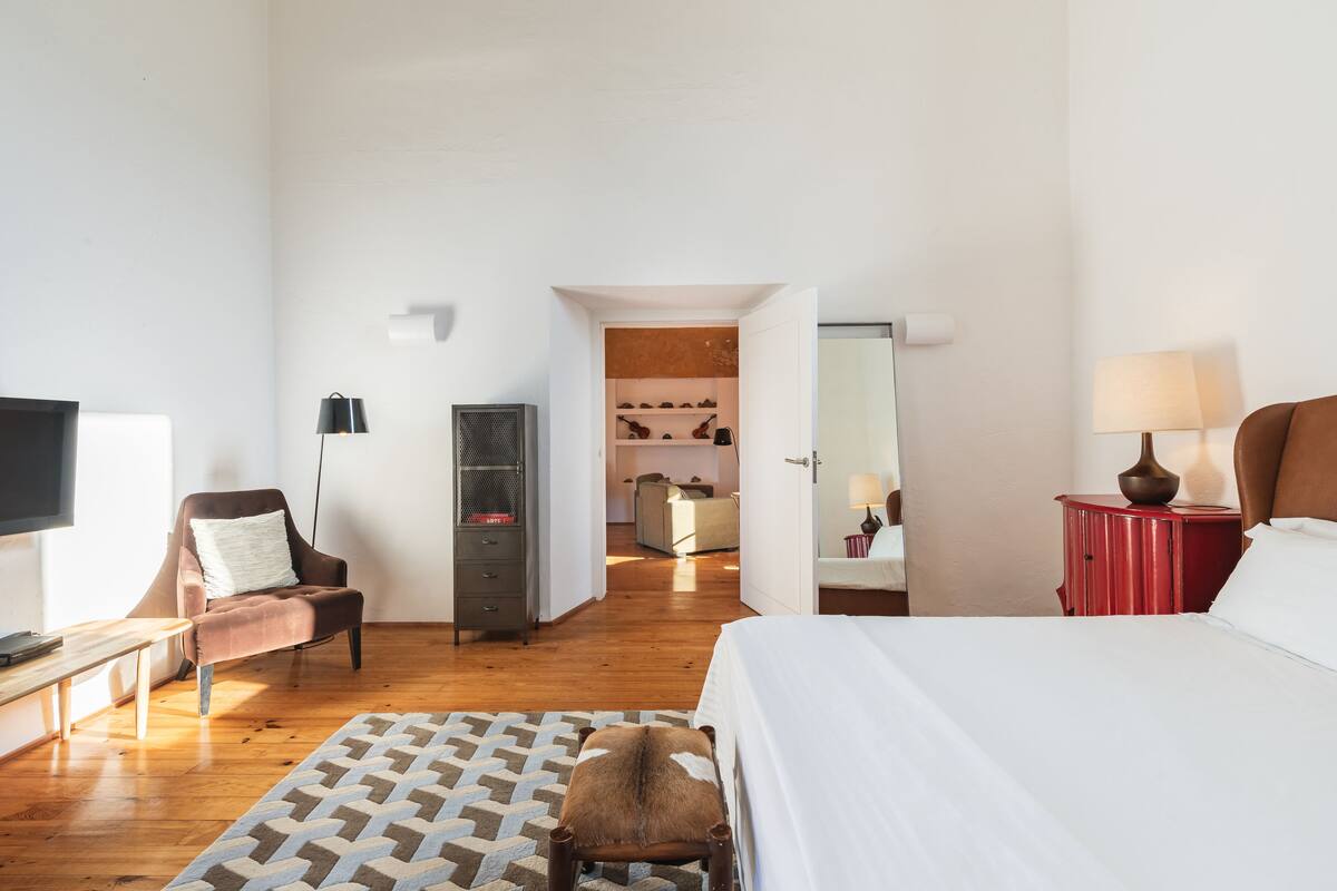A hotel room with a bed, a desk, a chair, a TV, a wardrobe, and a bathroom visible through an open door.