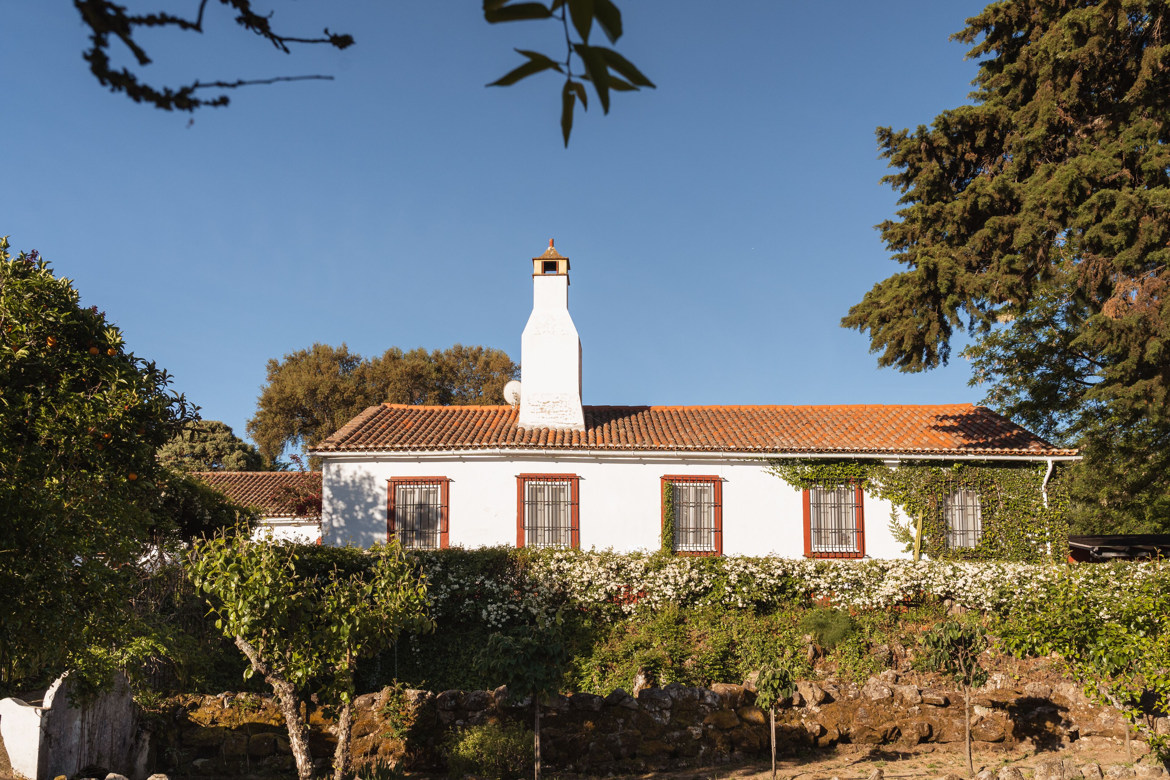 A white building with a red-tiled roof, surrounded by greenery and a stone wall.