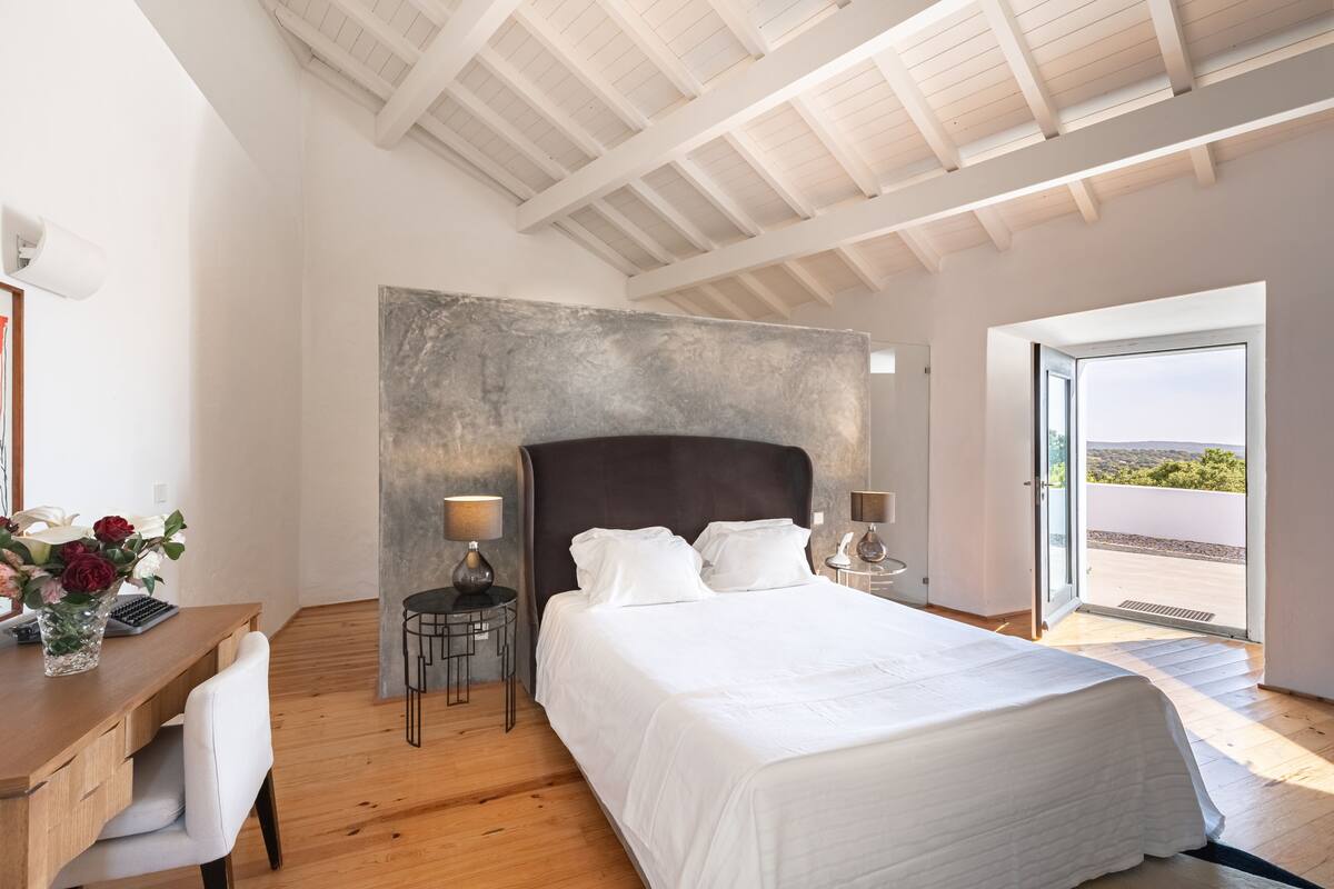 A bedroom with a large bed, a desk with a chair, a small table with a lamp, and a view through an open door.