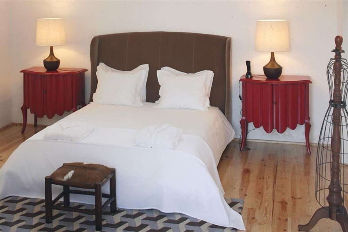 Una cama bien hecha con sábanas blancas, dos mesitas de noche con lámparas, un taburete pequeño y una alfombra estampada.