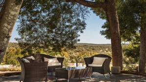 An outdoor seating area with wicker furniture and a stone table, surrounded by trees and hills in the background. - Imani Country House (Évora)
