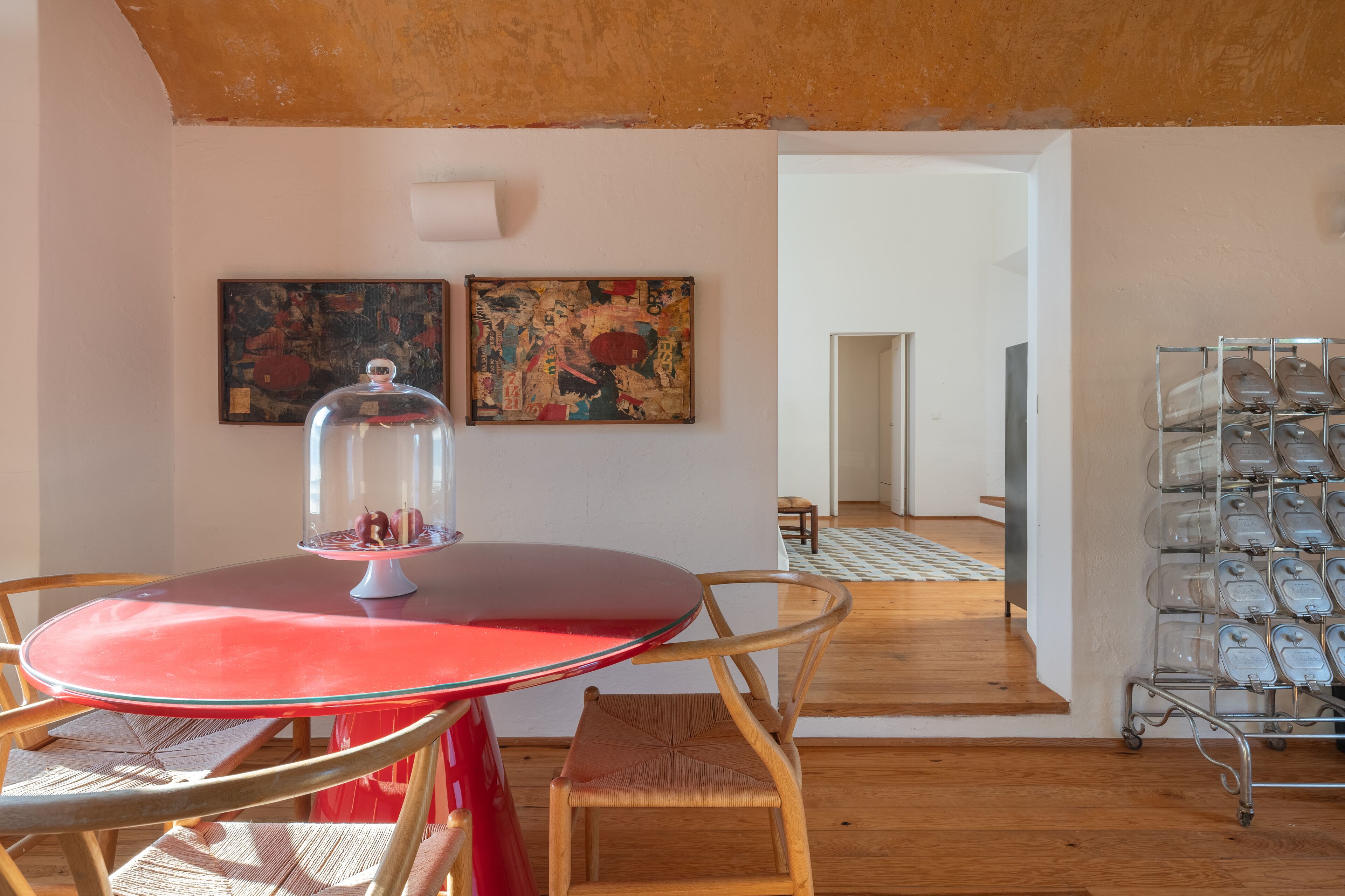 A dining area with a red table and wicker chairs, a glass display case with a dome, and two abstract paintings on the wall.