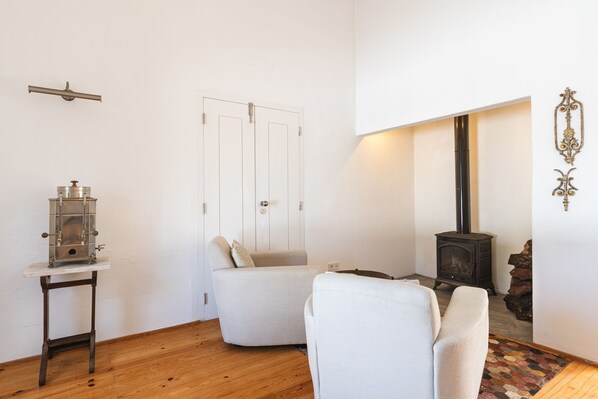 A room with a white wall, a wooden floor, a white armchair, a small table with a metal object, a door with a handle, and a wood-burning stove.