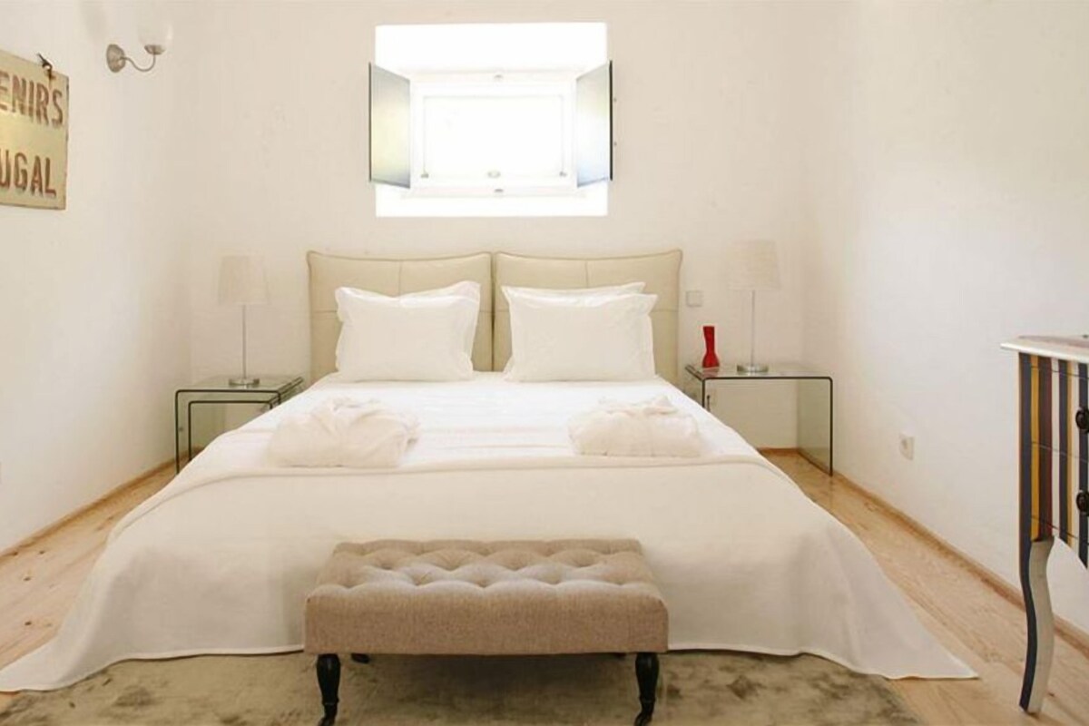 A bedroom with a bed, two bedside tables, a bench, a wall-mounted mirror, and a sign reading 'Souvenirs de Portugal'.