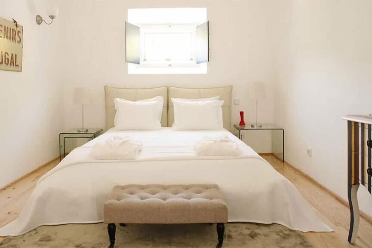 A bedroom with a bed, two bedside tables, a bench, a wall-mounted mirror, and a sign reading 'Souvenirs de Portugal'.