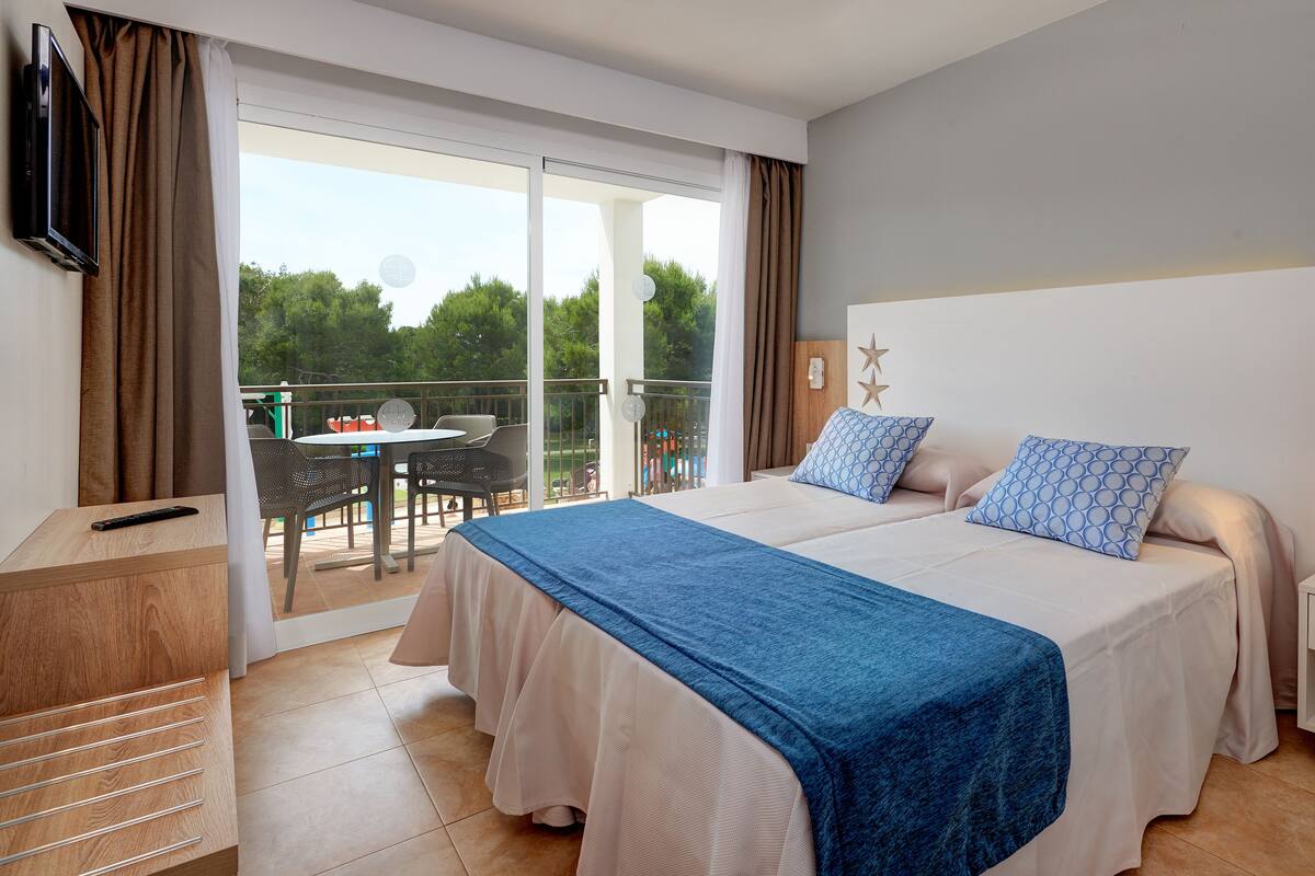 Un dormitorio con cama, televisor, balcón con mesa y sillas, y vista a árboles.