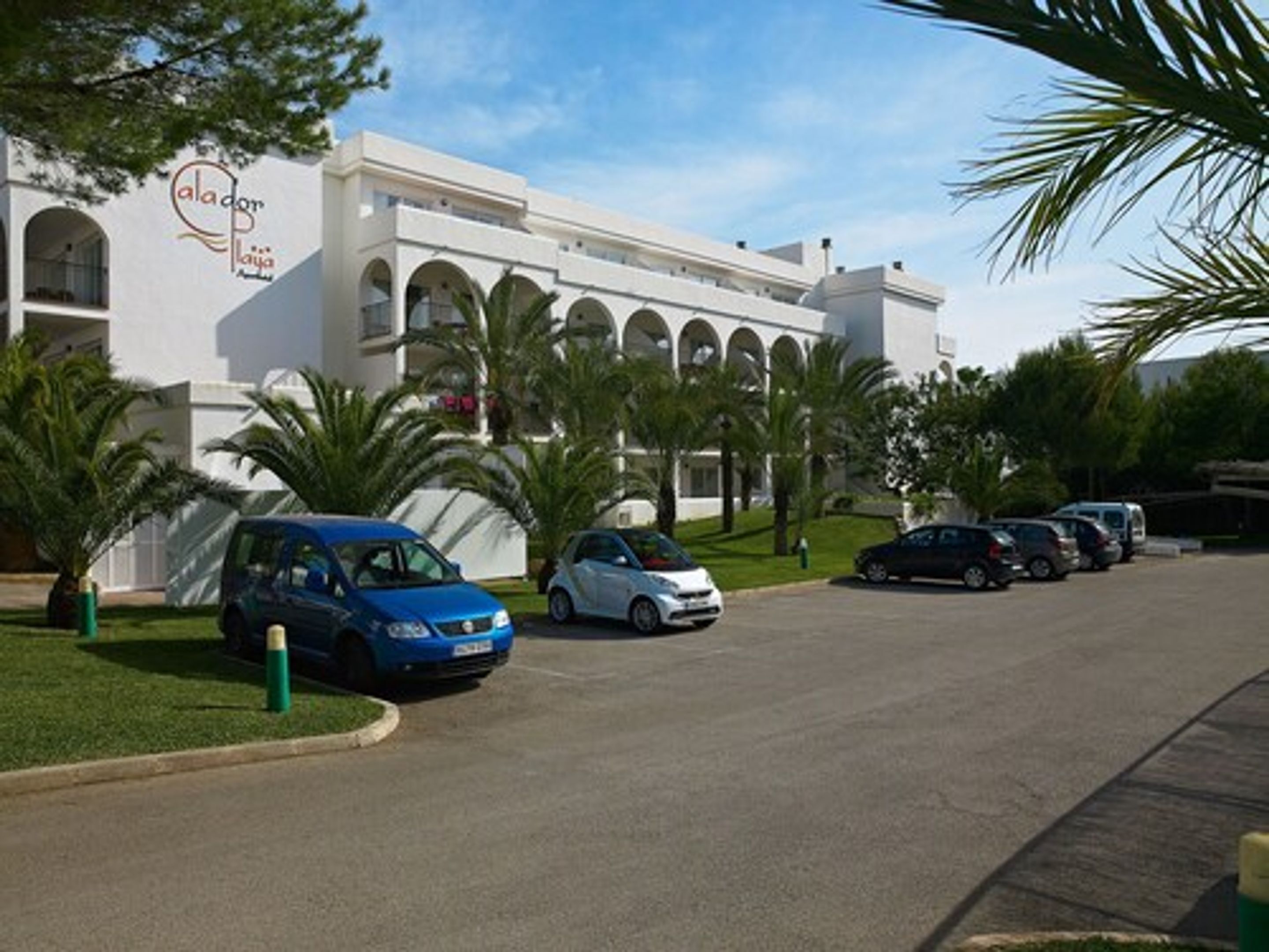 A modern building with a white facade, arched windows, and a logo on the side. There are several cars parked in front, and palm trees are visible.