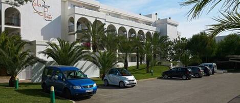 A modern building with a white facade, arched windows, and a logo on the side. There are several cars parked in front, and palm trees are visible.