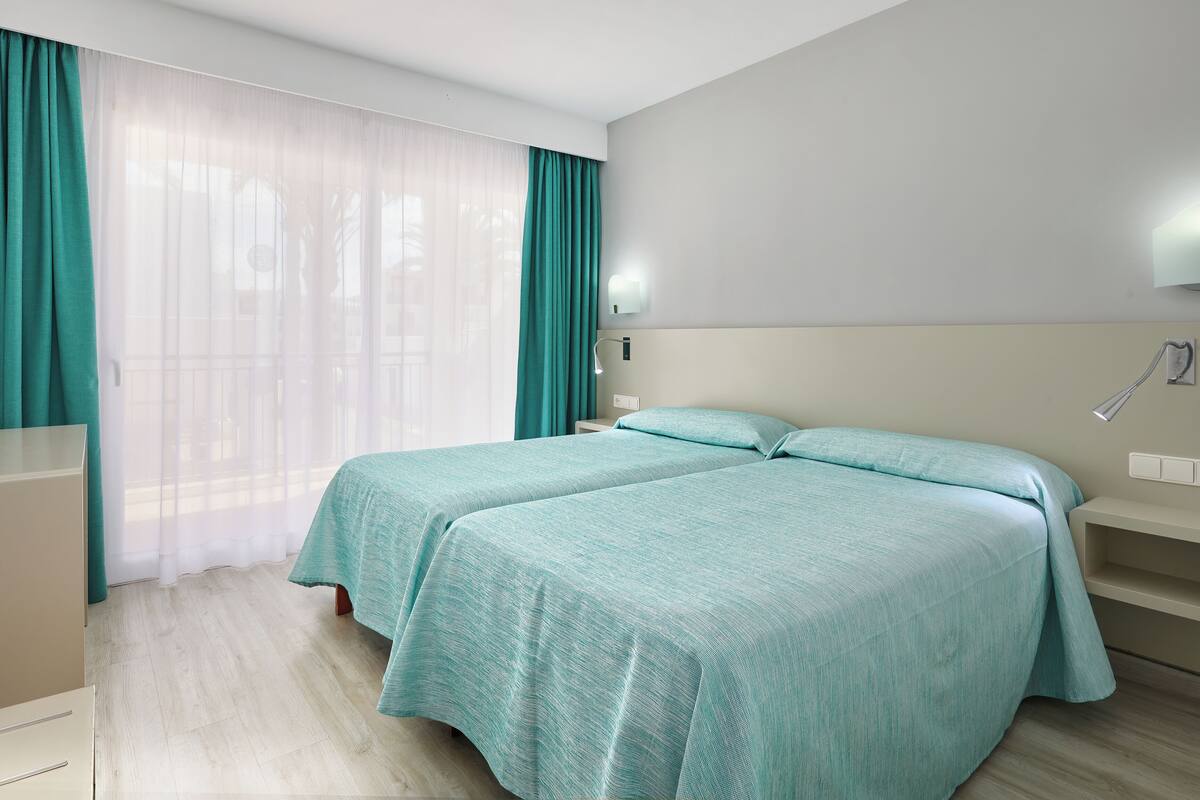 Habitación de hotel con dos camas, un ventanal grande con cortinas traslúcidas, una mesita de noche y una lámpara de pared.