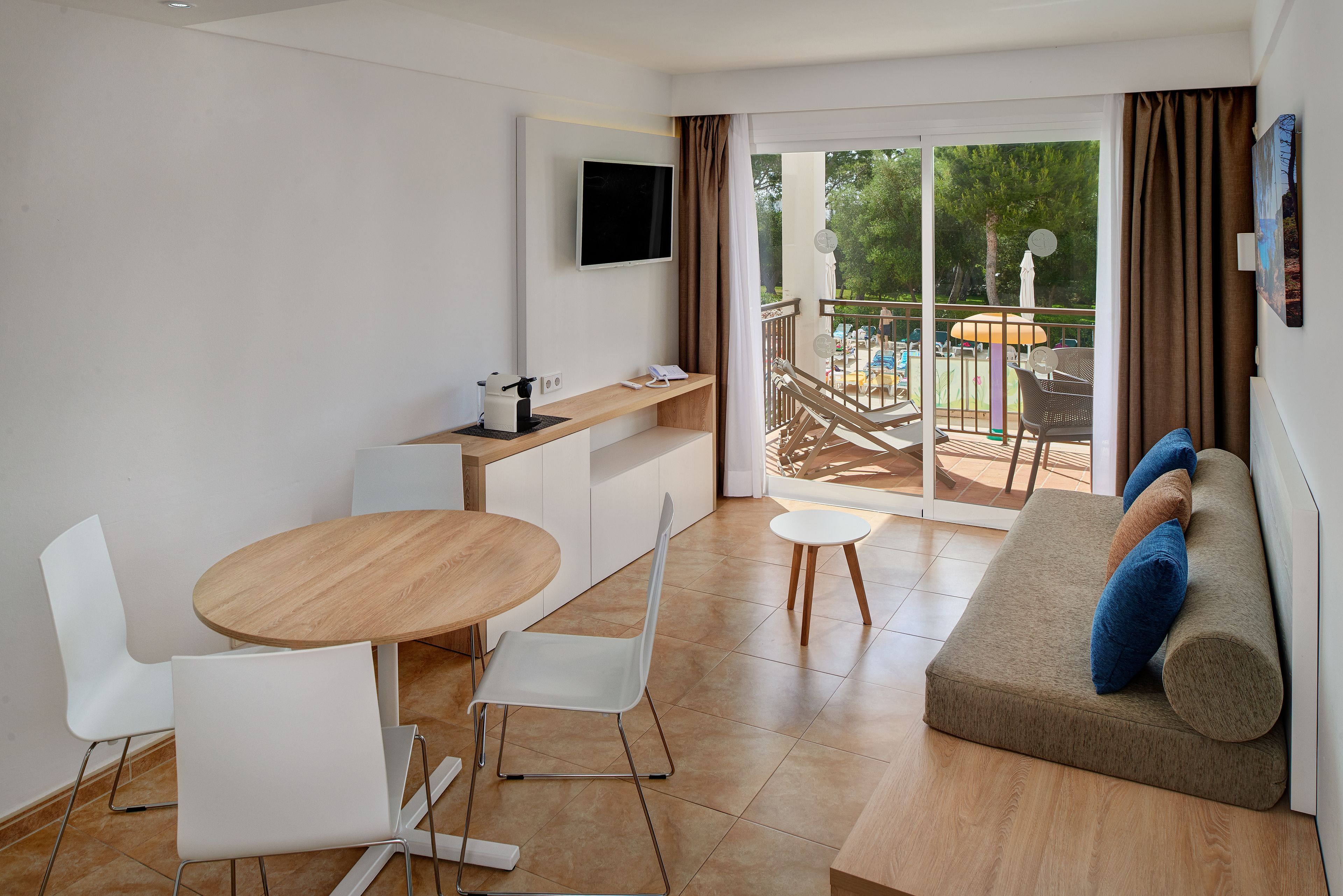 A modern, well-lit room with a dining table, chairs, a sofa, and a TV on the wall.