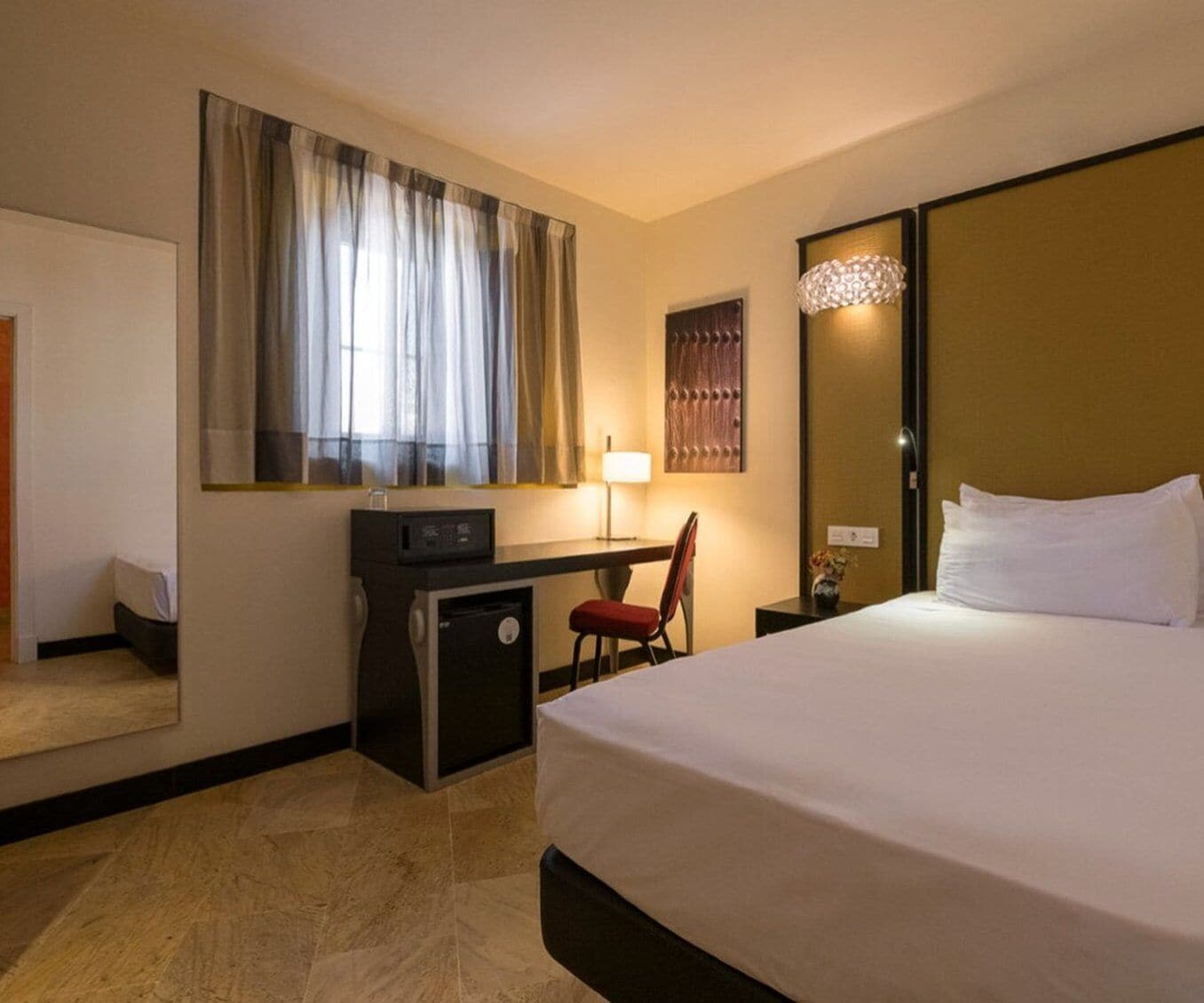 A hotel room with a bed, a desk with a chair, a lamp, and a large window with curtains.