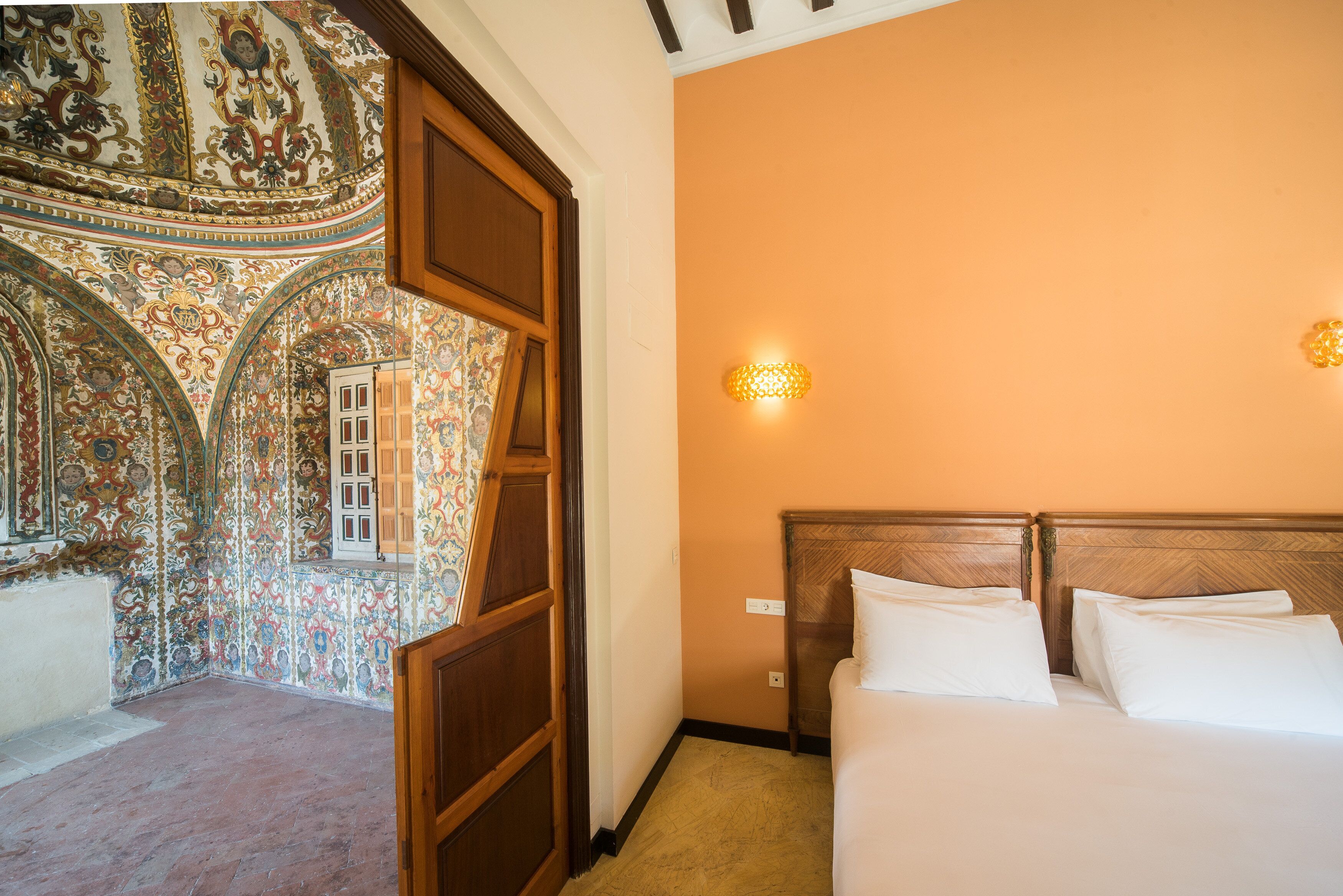 A hotel room with a large bed, a wooden headboard, a decorative wall with intricate patterns, and a small wall-mounted light fixture.