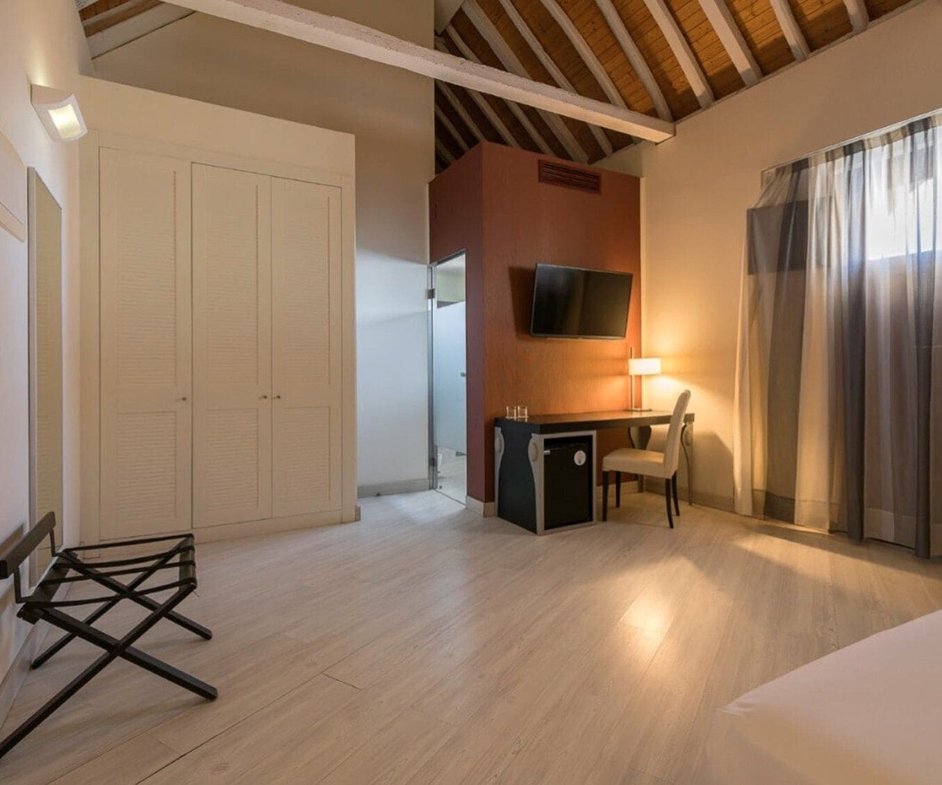 A hotel room with a wooden ceiling, a desk with a chair, a TV mounted on the wall, and a large window with curtains.