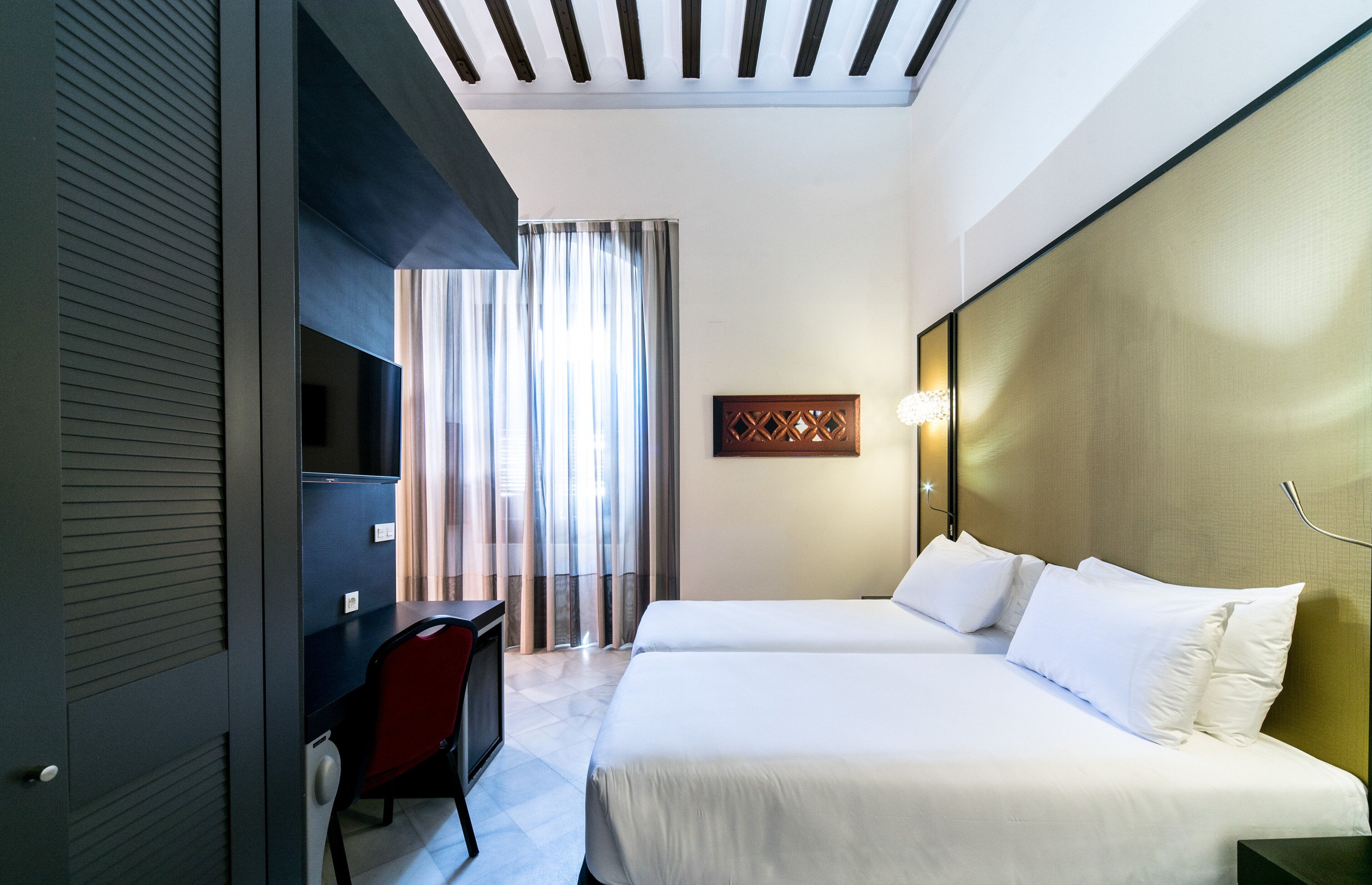 Une chambre d’hôtel avec deux lits, un bureau avec une chaise, une télévision et une grande fenêtre avec des rideaux.