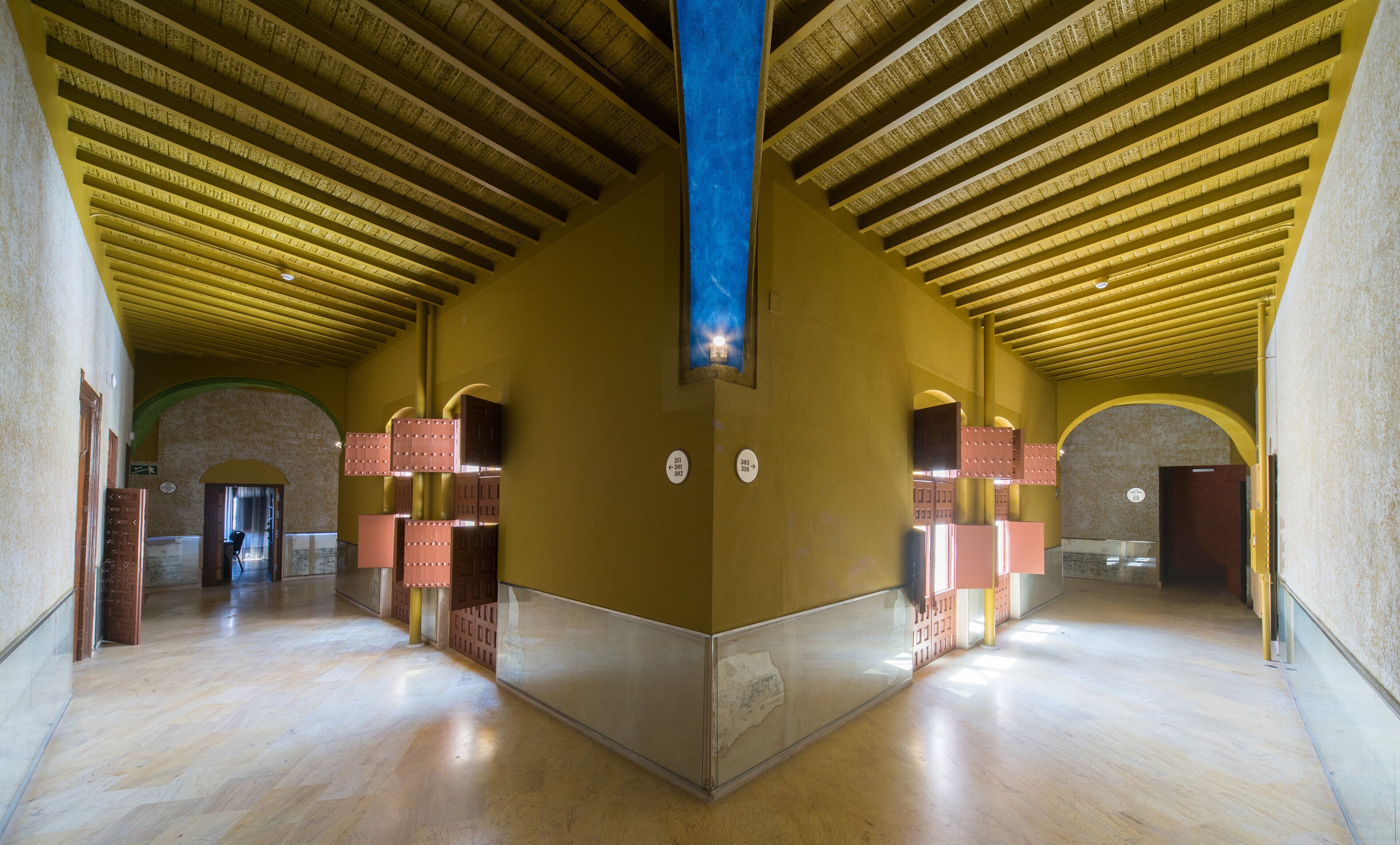 Un interior espacioso con techos altos, una viga azul central y varias puertas que dan a diferentes espacios.