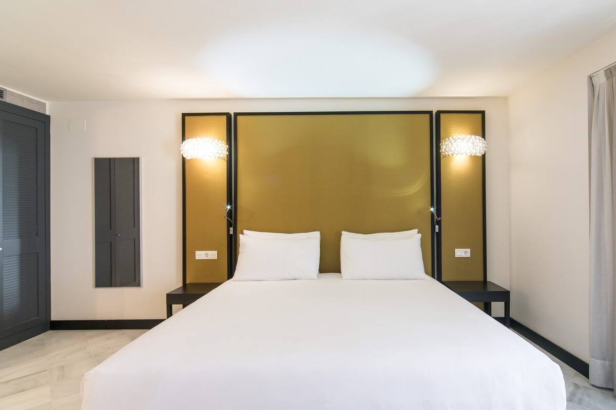 Una habitación de hotel con una cama grande, dos mesitas de noche, un cabecero con iluminación integrada y una ventana con cortinas.